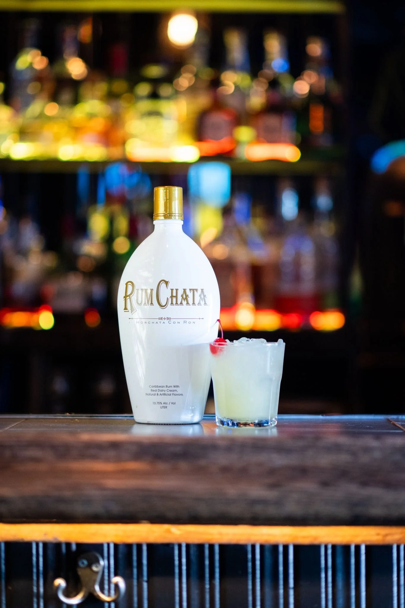 A bottle of RumChata and a cocktail garnished with a cherry and pineapple, placed on a wooden bar counter with blurred colorful bottles in the background.
