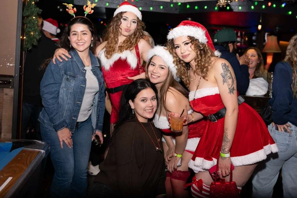 Group of women celebrating Christmas, some dressed in holiday costumes and Santa hats, holding drinks in a festively decorated setting.