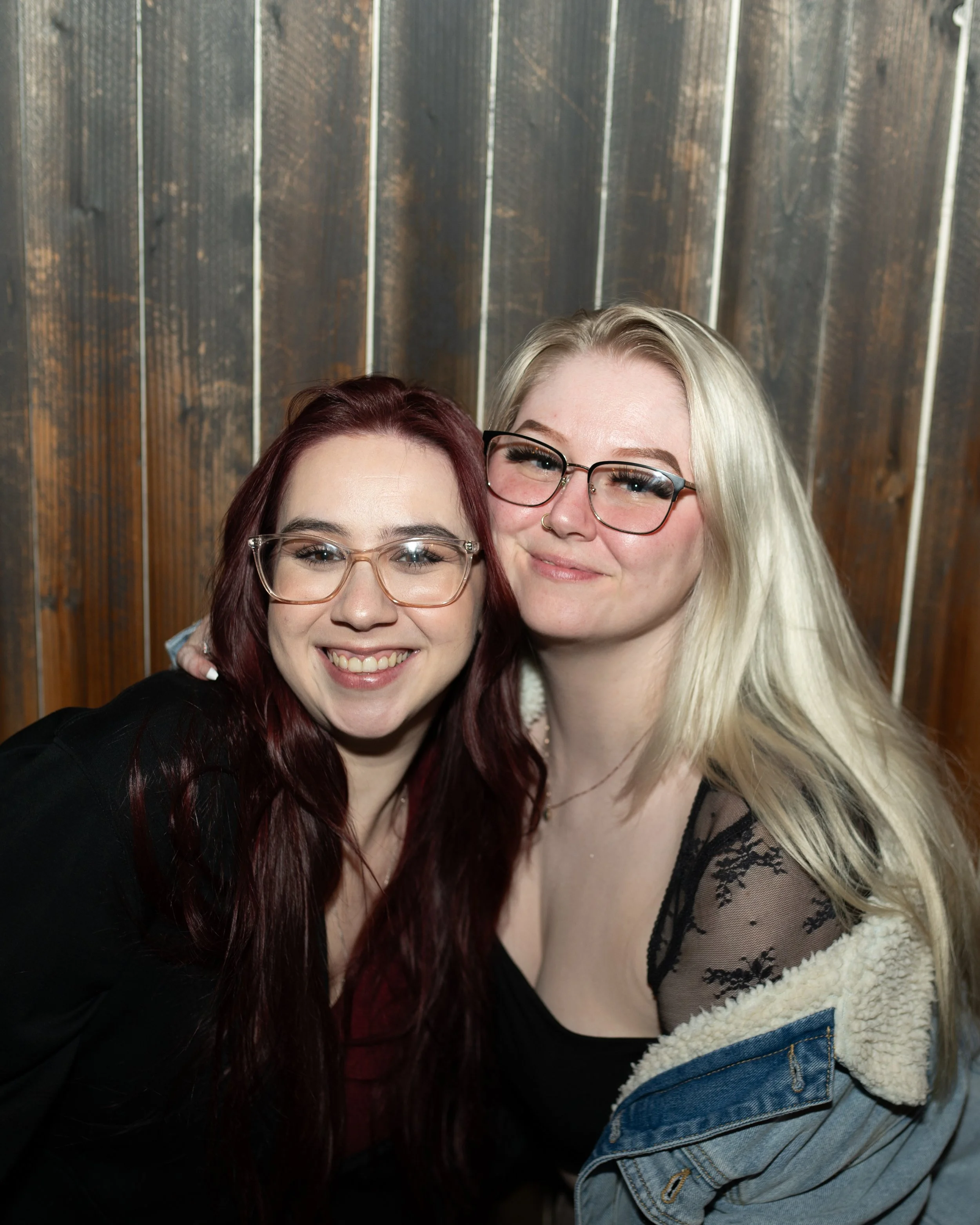 Two women with glasses smiling and posing together in front of a wooden wall