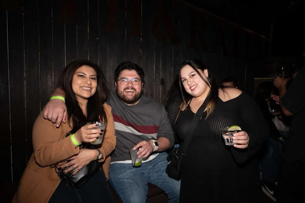 Three friends at a bar enjoying drinks, smiling and posing for the camera, with a dark background and other people in the distance.