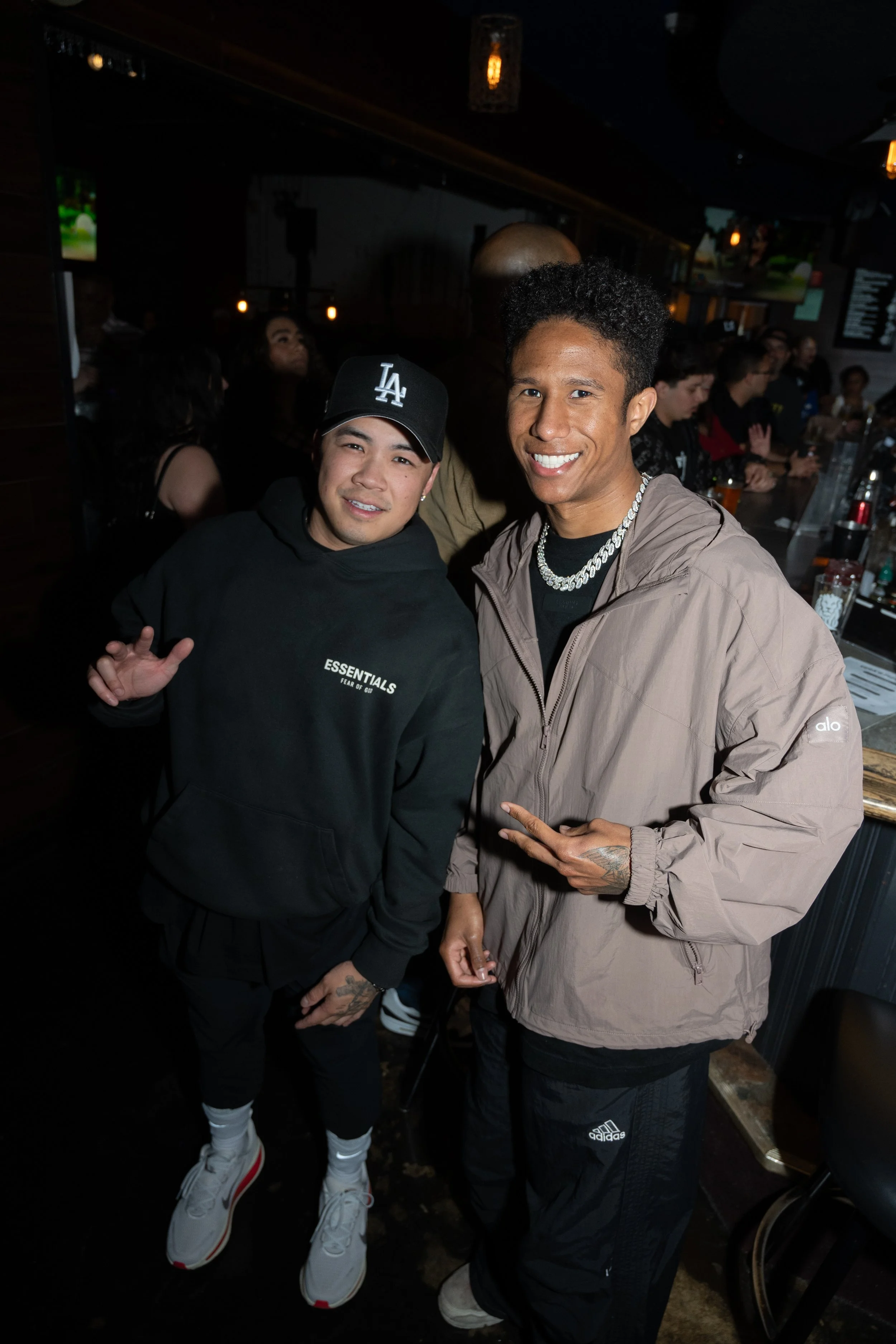 Two men posing indoors at a lively event, one wearing a black cap and hoodie, the other in a brown jacket and jewelry, both smiling.