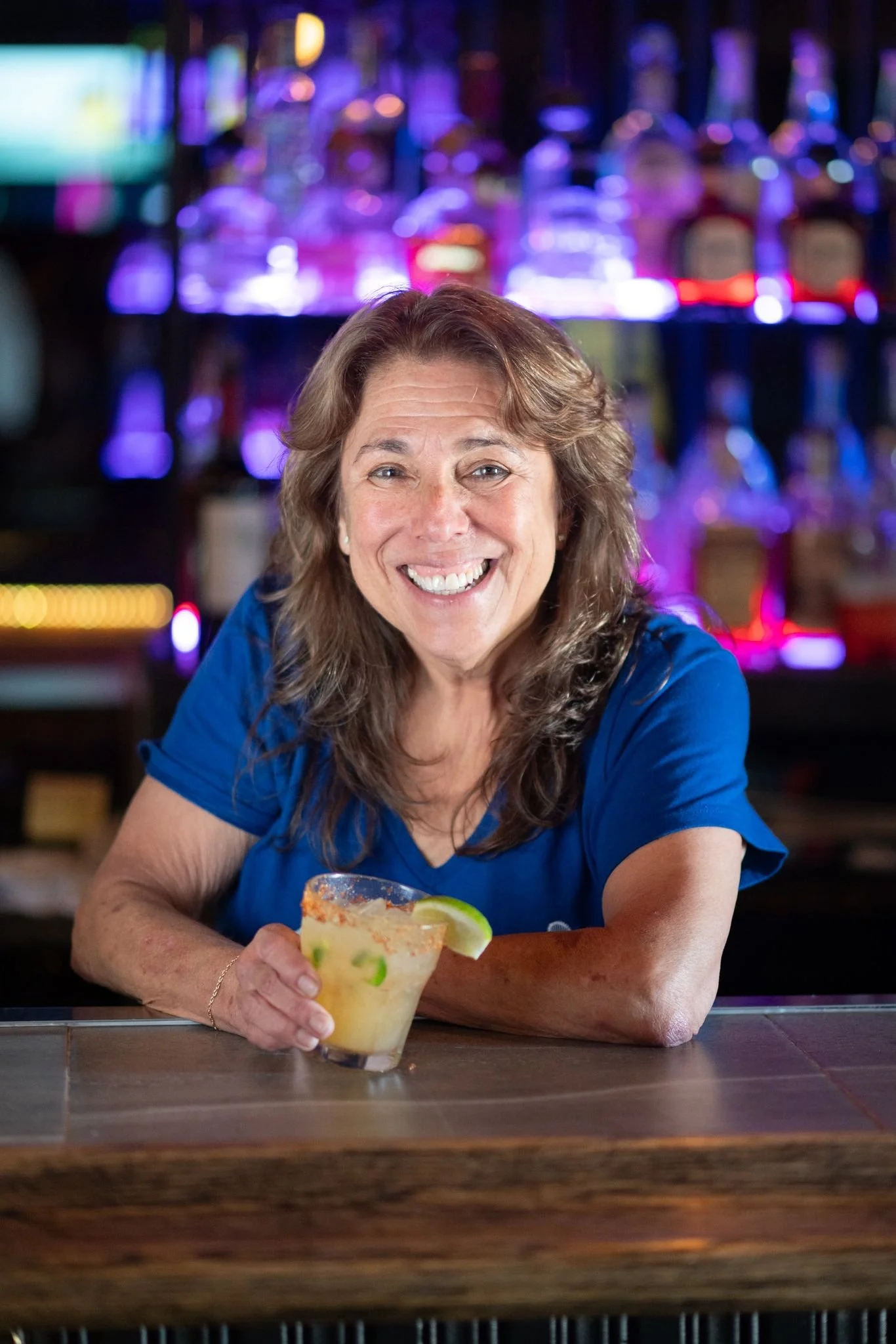 A smiling woman in a blue shirt sitting at a bar with a margarita in her hand.