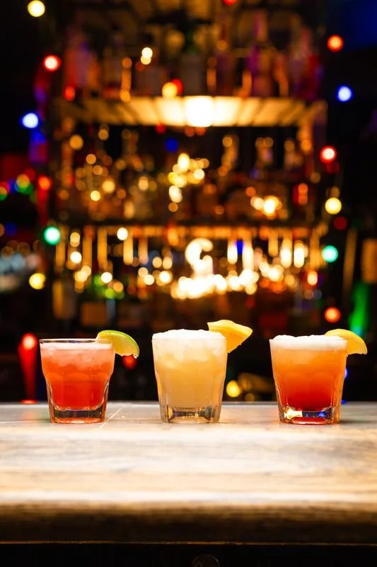 Three cocktails on a wooden bar counter, each garnished with fruit, with colorful lights and blurred bar shelves in the background.