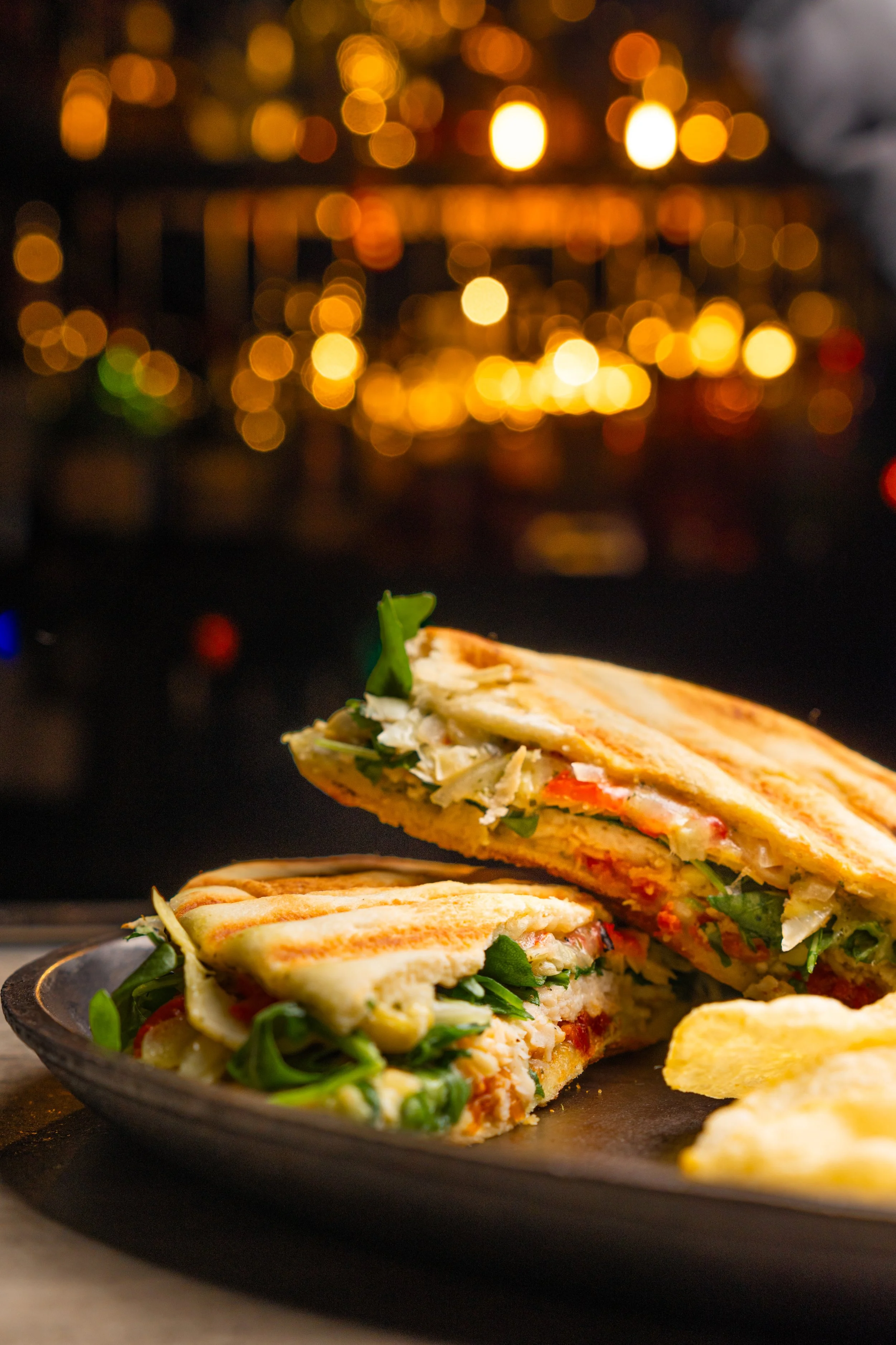 Grilled panini sandwich with vegetables on a black plate against a blurred background of warm lights.