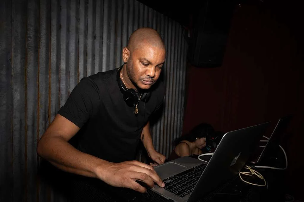 A man wearing a black shirt using a laptop, with headphones around his neck, in a dimly lit room with a corrugated metal wall in the background.