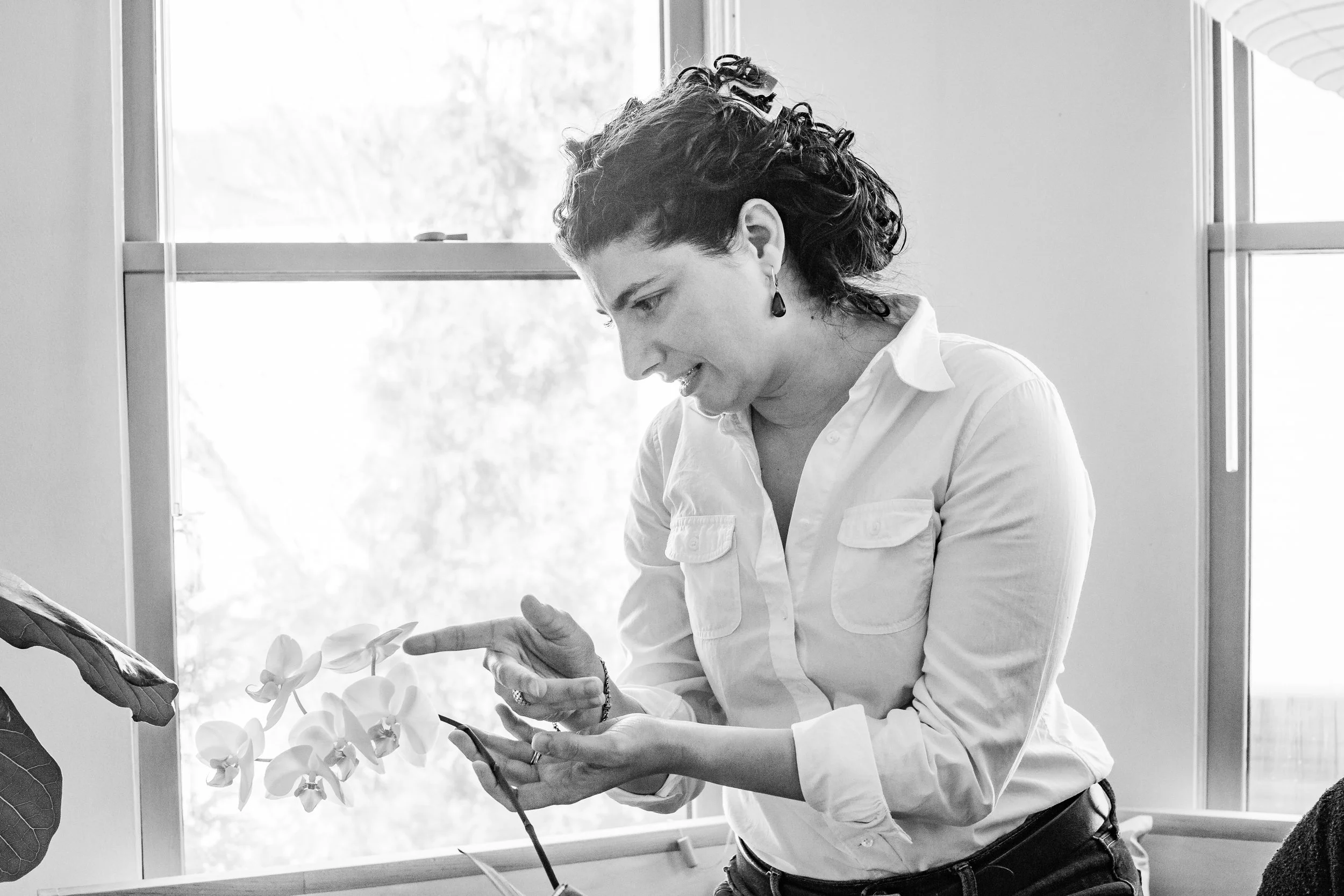 A woman with short, curly hair and earrings looks at her smartphone, pointing at a flower plant with her finger inside a room with large windows.
