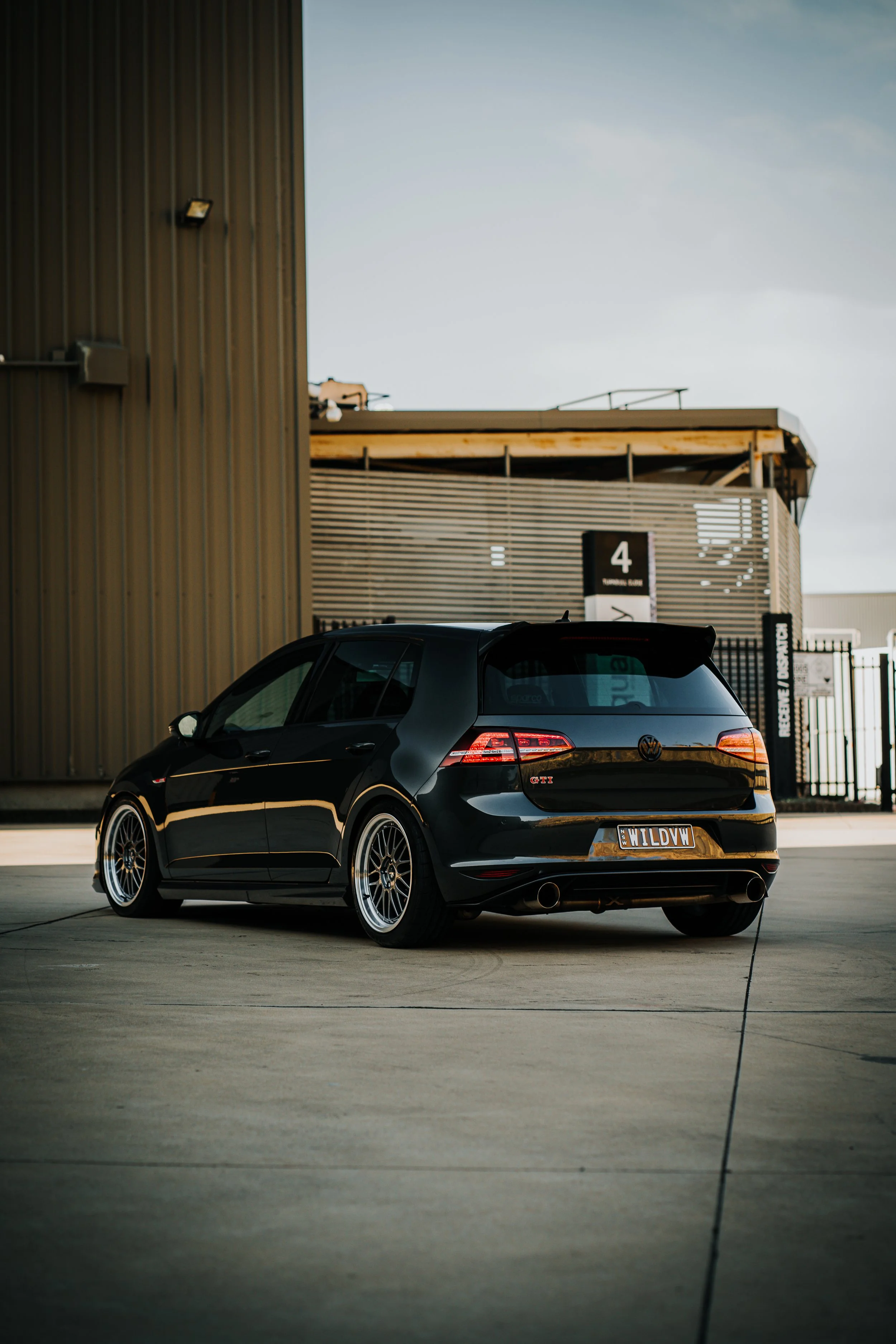 A black Volkswagen Golf GTI parked outside in an industrial area. The car has aftermarket wheels and a custom license plate reading 'WILD VW'.