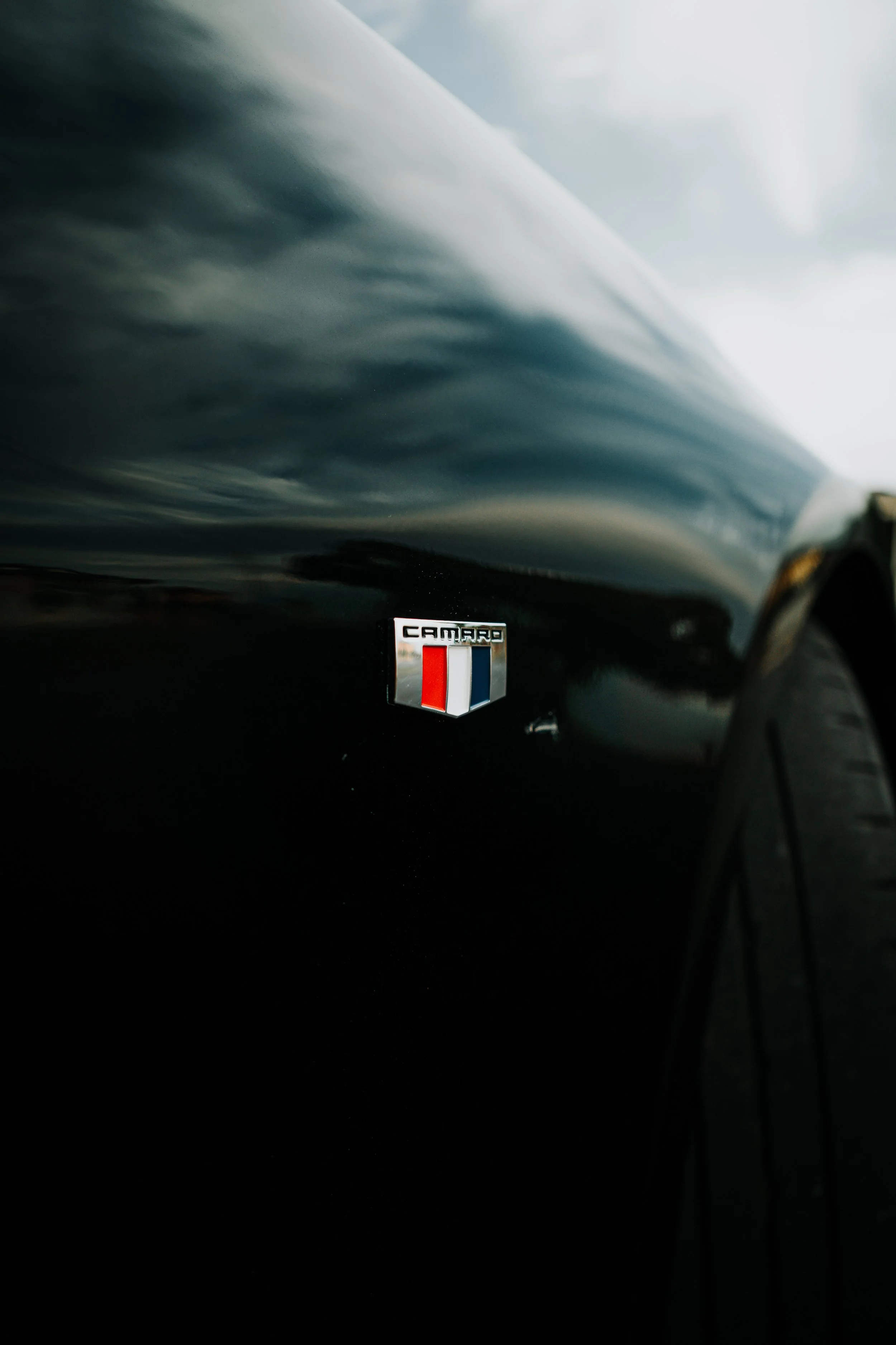 Close-up photo of a black Chevrolet Camaro with the Camaro badge on the side, showing part of the front wheel and the car's reflective surface.