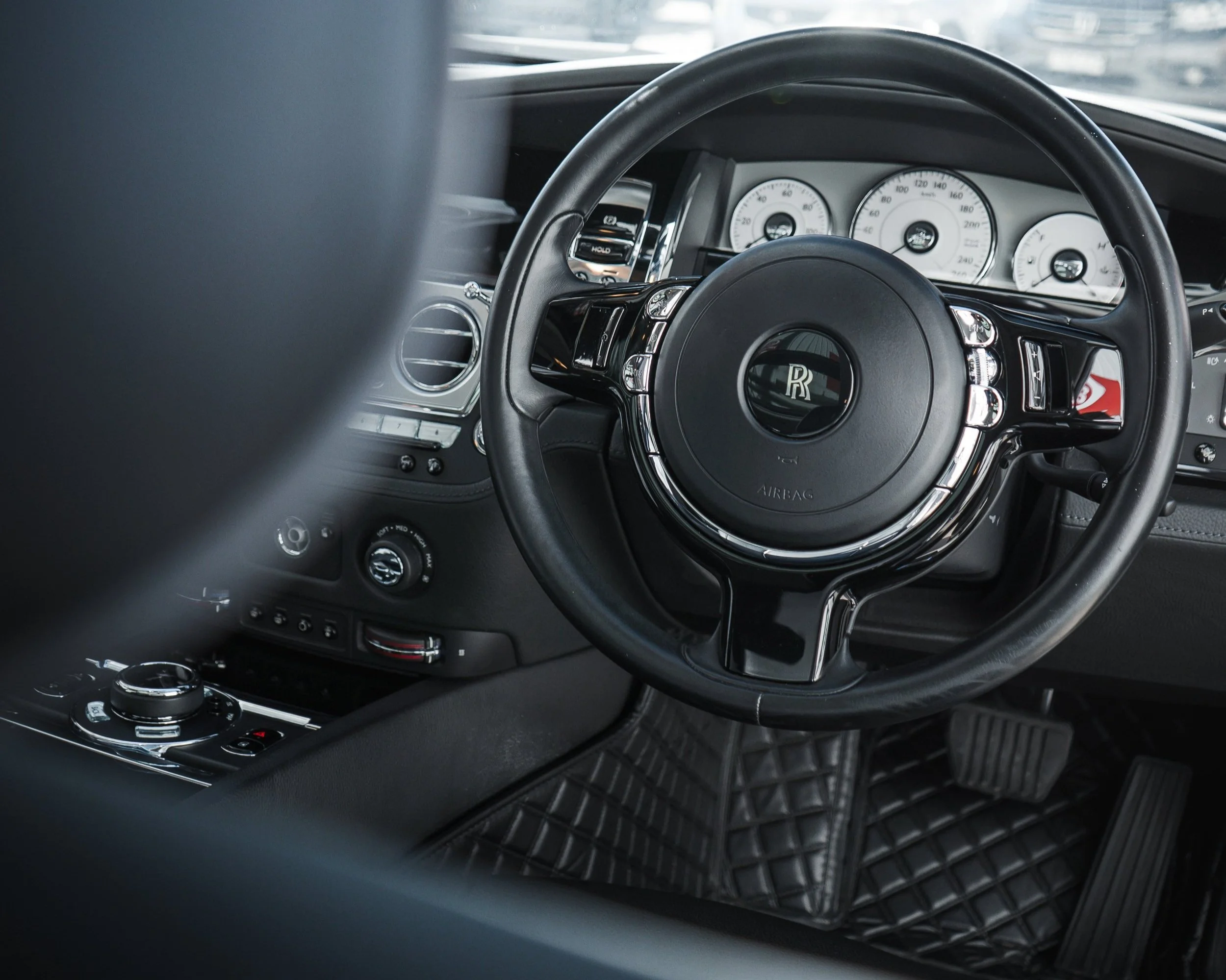 Interior of a luxury car showcasing the steering wheel with Rolls-Royce emblem, dashboard with speedometer and gauges, control knobs, and black quilted floor mats.