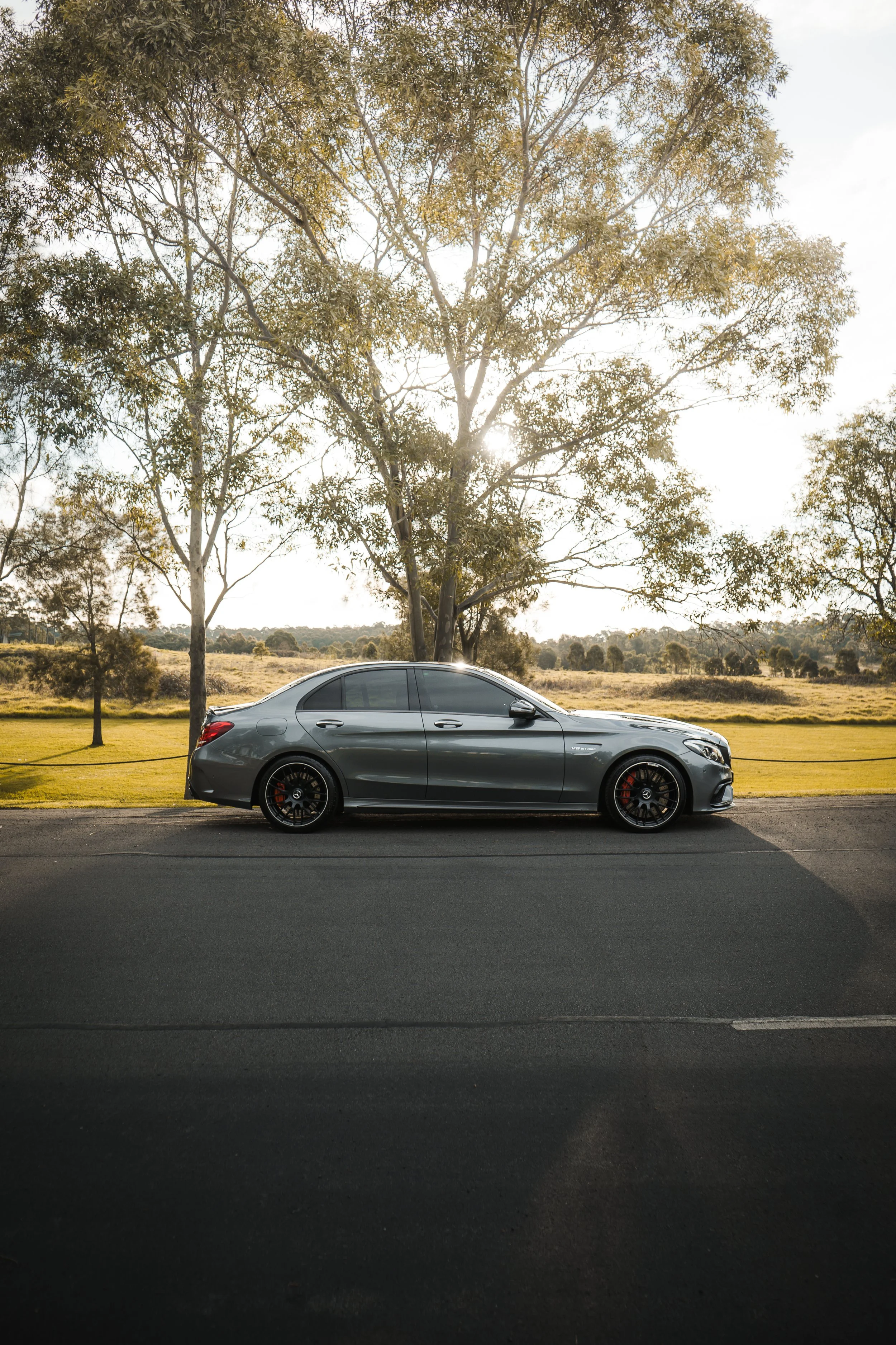 A gray luxury sedan parked on the side of a paved road, with a backdrop of grassy fields, trees, and a bright sun shining through the branches.