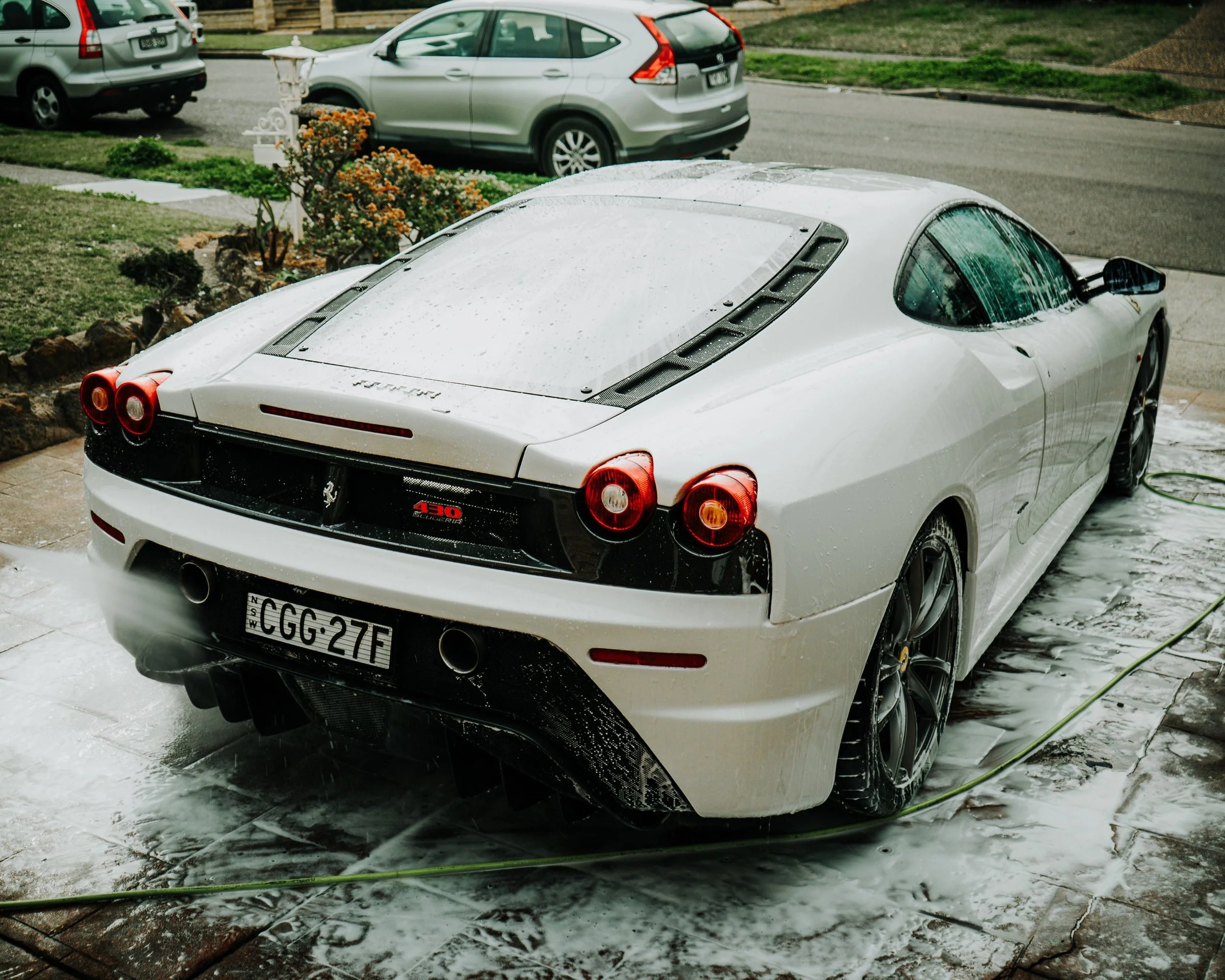 White Ferrari 430 Scuderia being washed on a driveway with soap and water, with a garden and parked cars in the background.