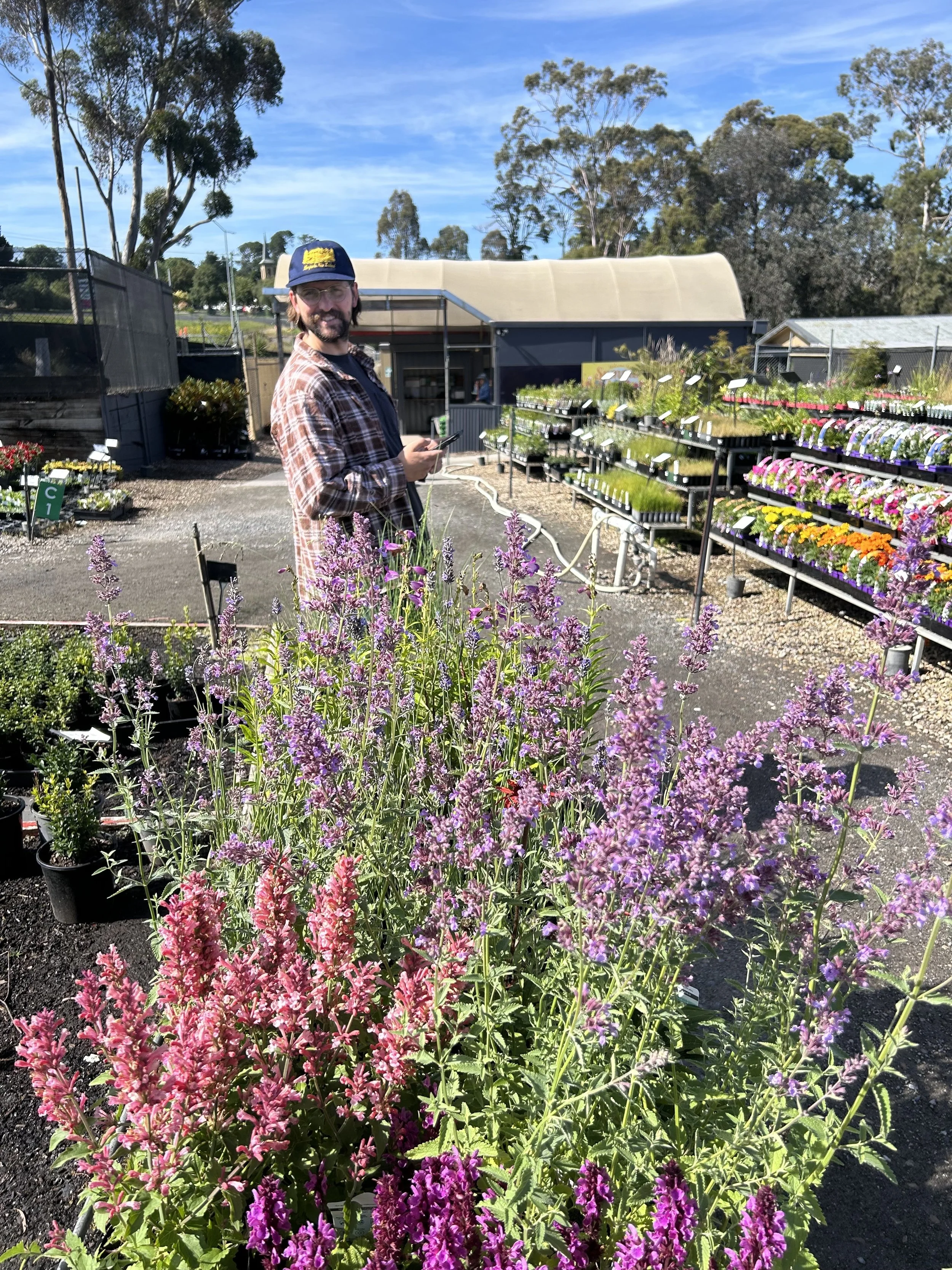 Man smiling in a garden center with colorful flowering plants in the foreground and shelves of other plants in the background.