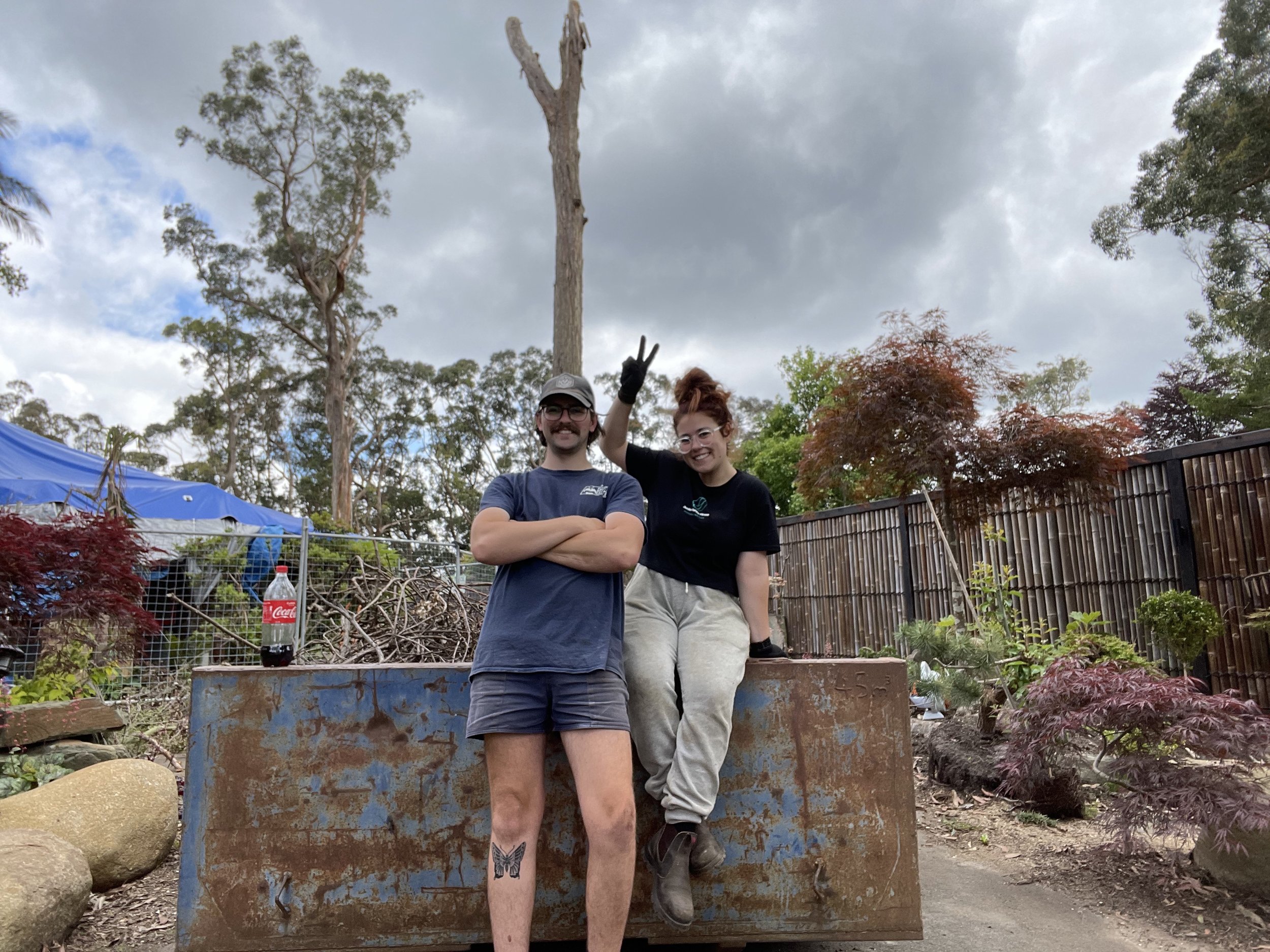 Two people, a man and a woman, posing outdoors on a rusty metal platform in a garden, with trees, bushes, and a cloudy sky in the background. The woman is sitting on the platform, raising a peace sign, and the man is standing with crossed arms, smiling.