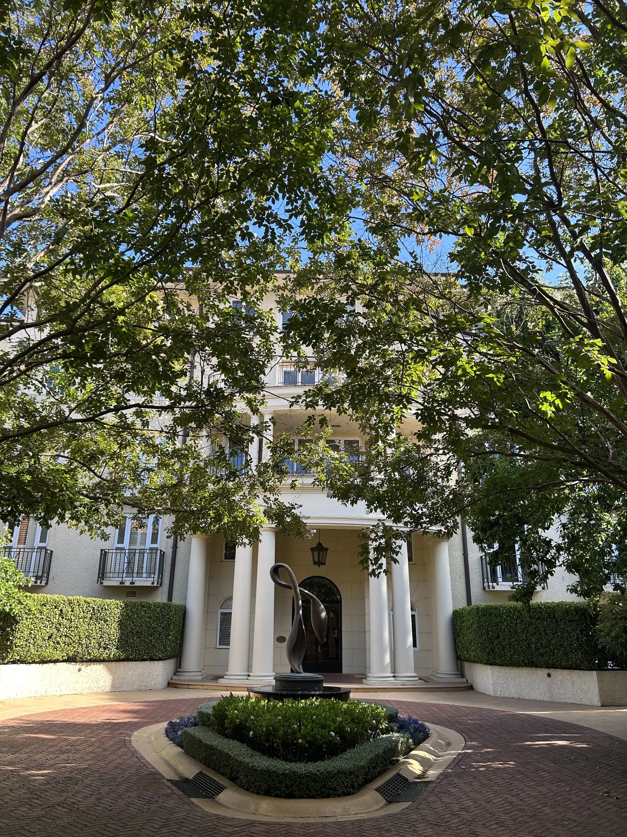Front view of a multi-story residential building with columns, balcony railings, and a curved driveway. There's a modern sculpture in the center of a flowerbed, surrounded by greenery and trees, under a partly cloudy sky.
