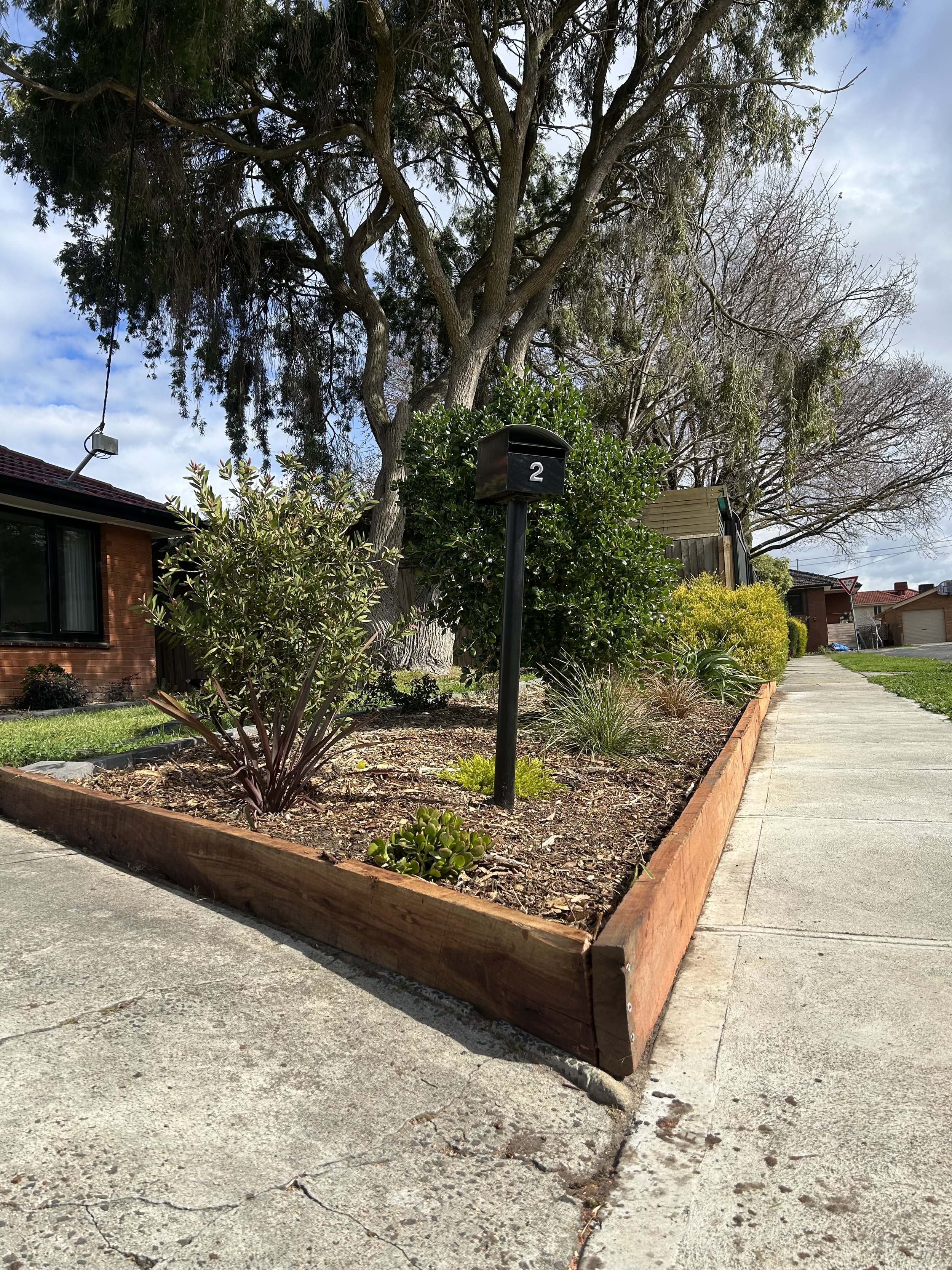 A sidewalk runs along a landscaped yard with various bushes and trees, including a large tree with sparse branches. A mailbox marked with the number 2 is situated among the bushes. There are houses and a street in the background under a partly cloudy