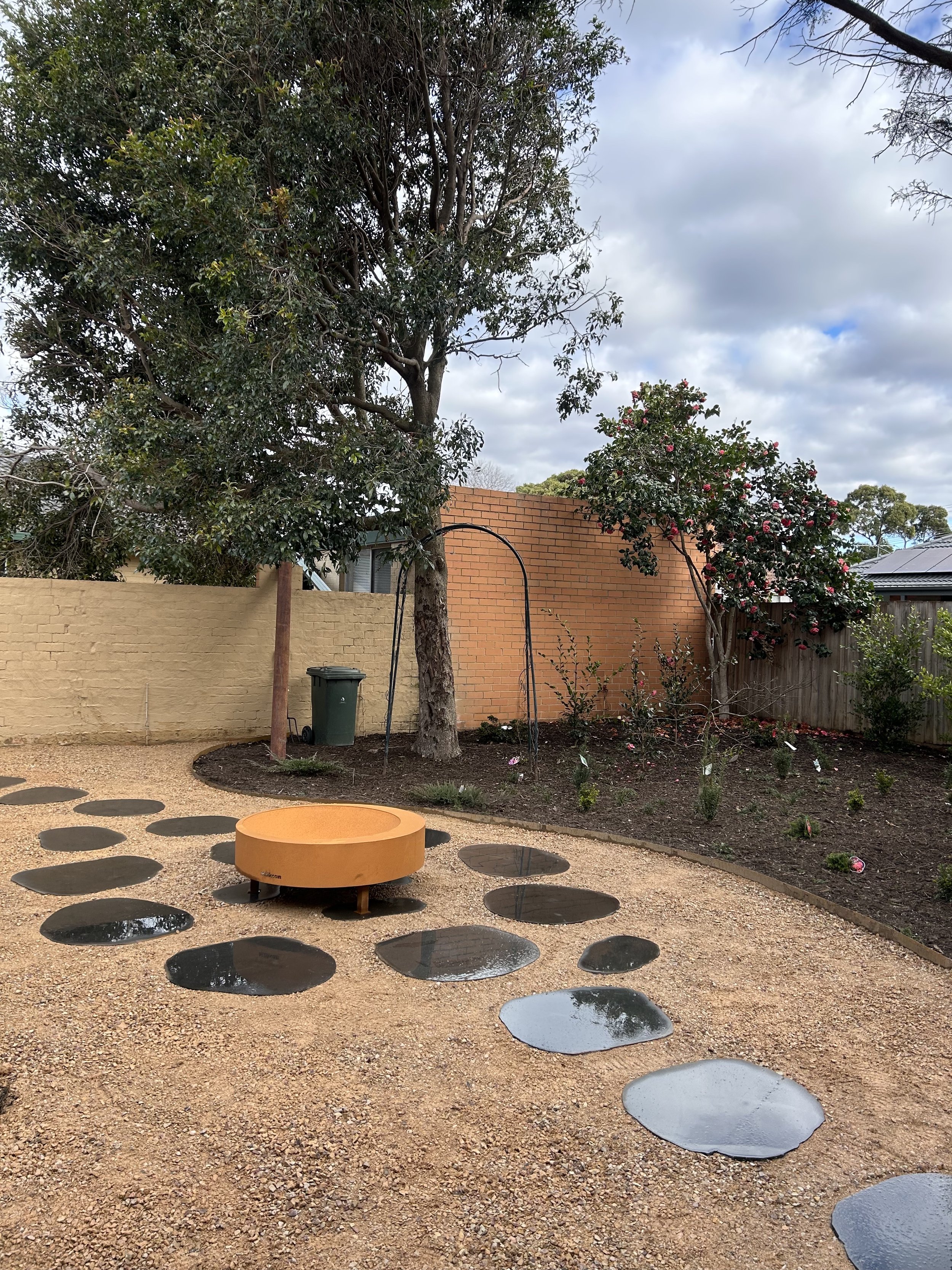 Backyard garden featuring a large tree, a flowering shrub, a weathered brick wall, and a water feature with stepping stones on a gravel surface.