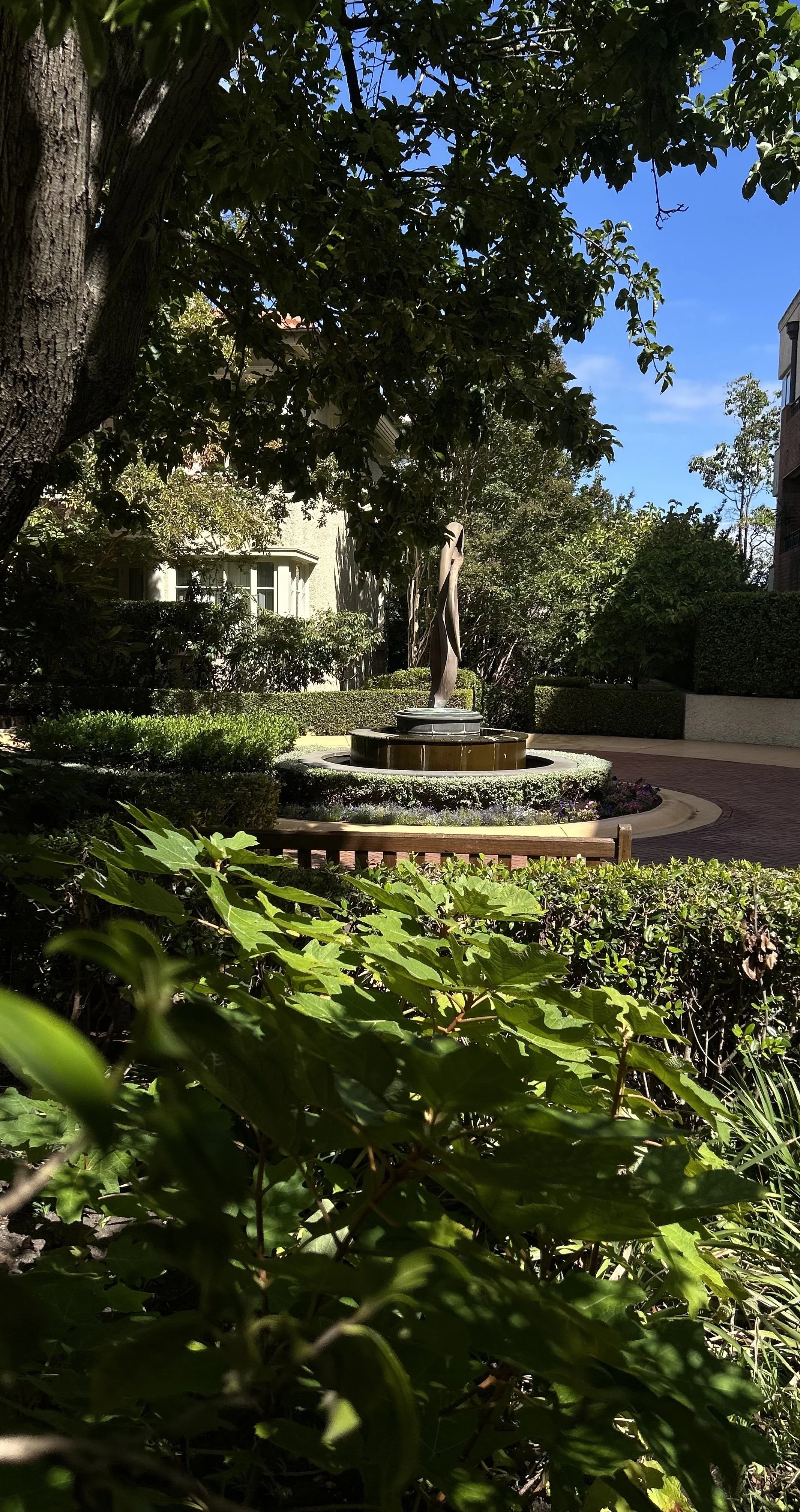 A small fountain with a bronze sculpture in a landscaped courtyard, surrounded by green bushes, trees, and a clear blue sky.