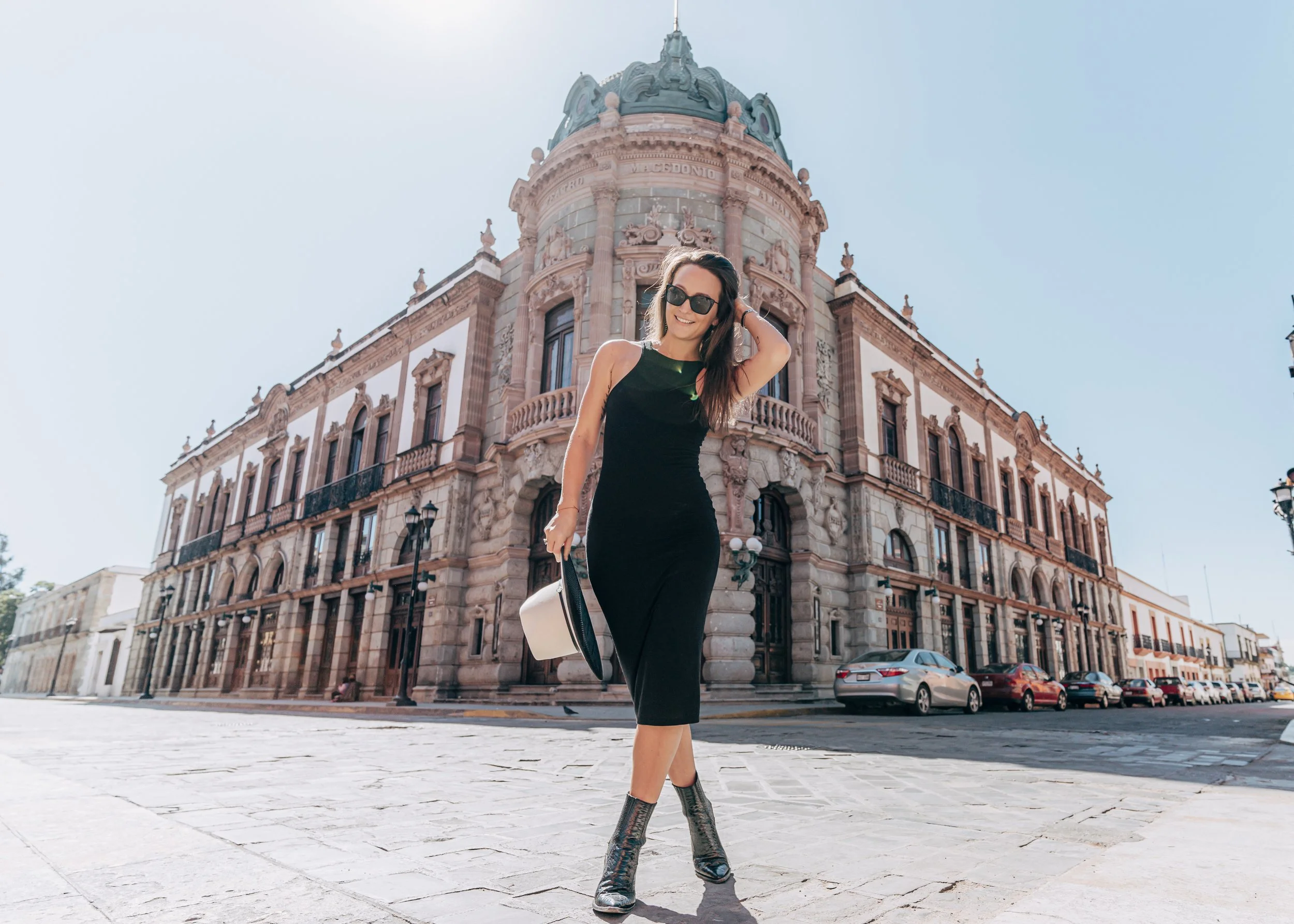 Mujer con vestido negro y sombrero en una plaza frente a un edificio histórico con arquitectura elaborada.