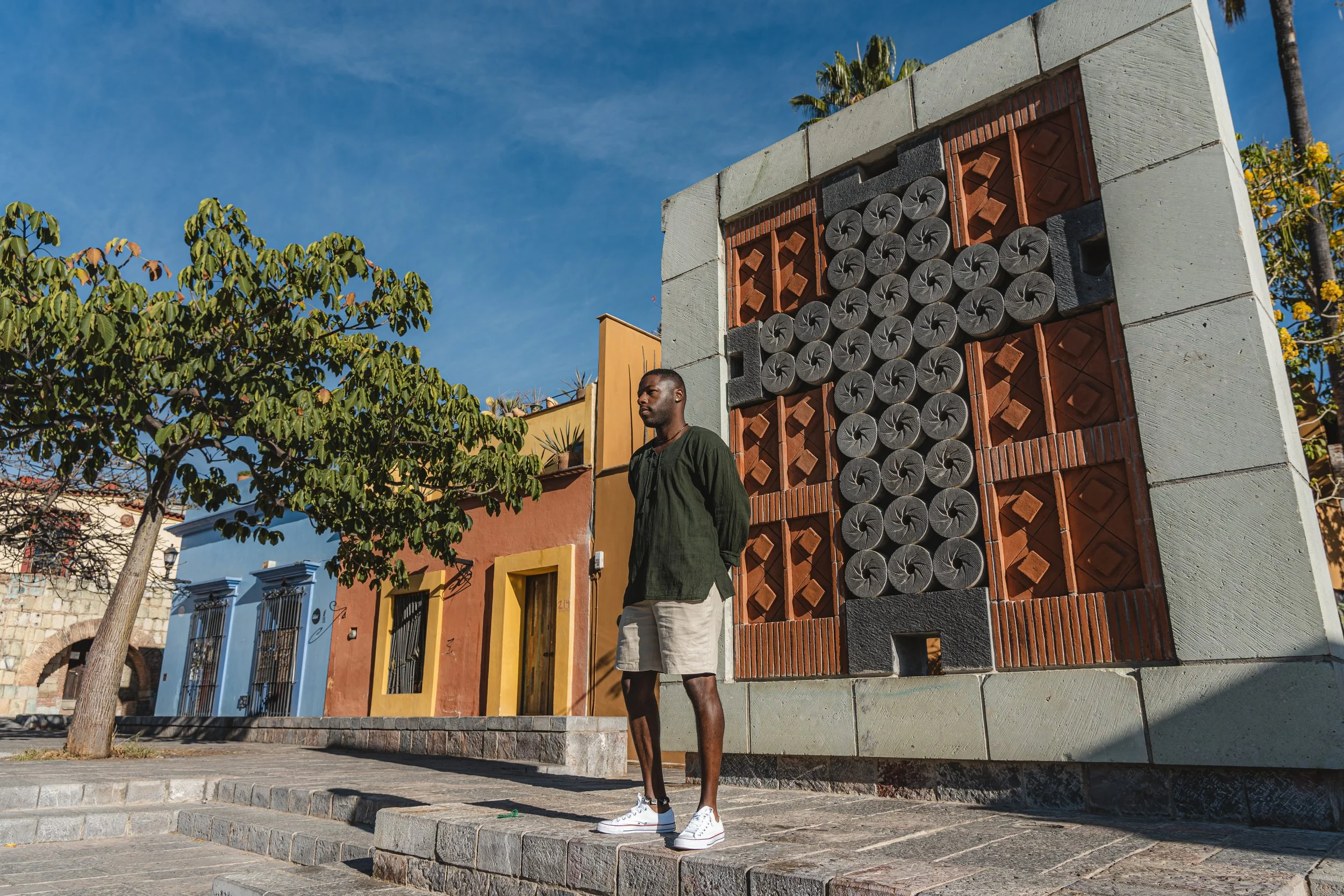 Persona de pie junto a una escultura de diseño geométrico en una plaza, con casas coloridas al fondo y un árbol a la izquierda.