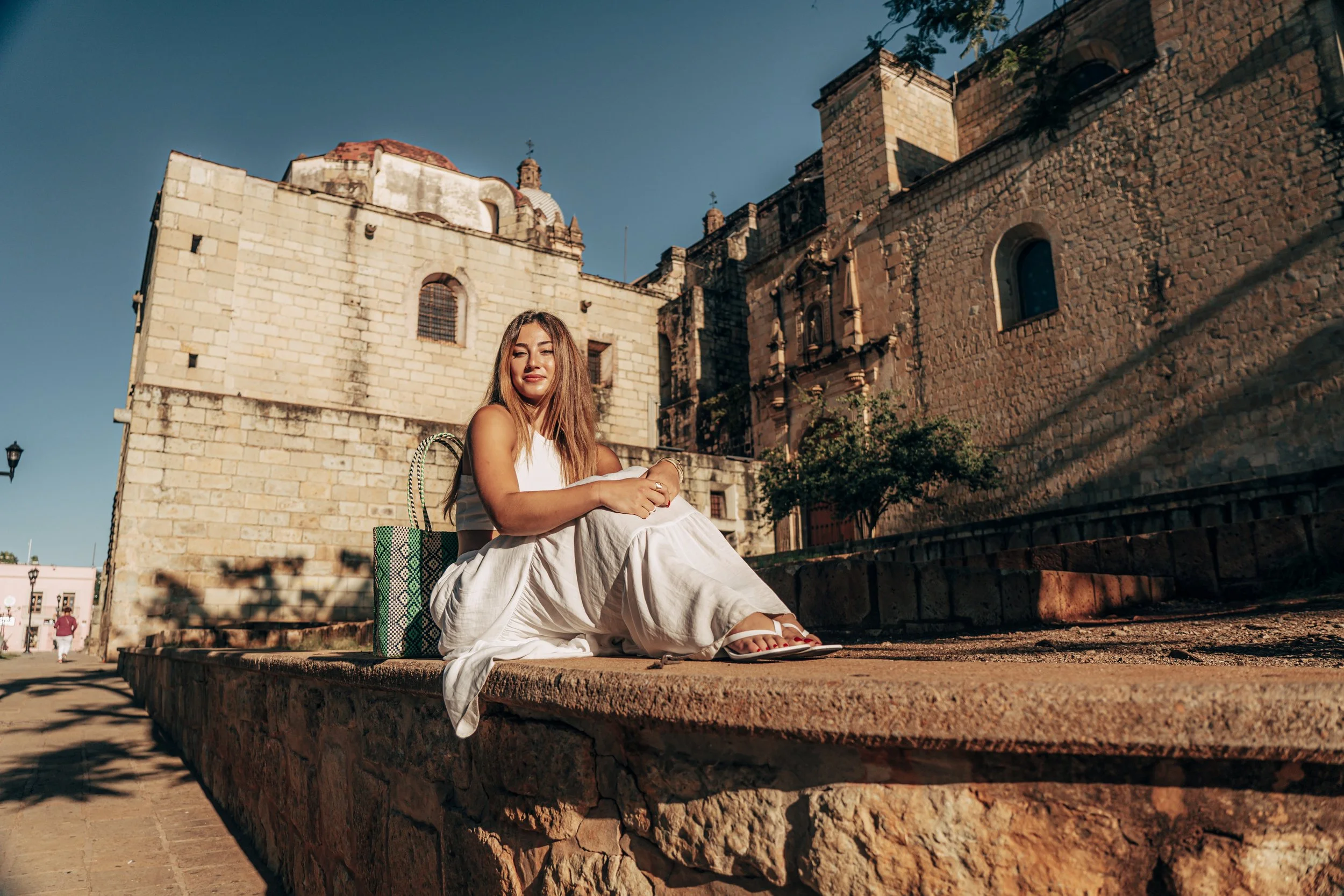 Mujer sentada en una pared de piedra en un entorno arquitectónico colonial, con un edificio histórico de fondo y cielo despejado.