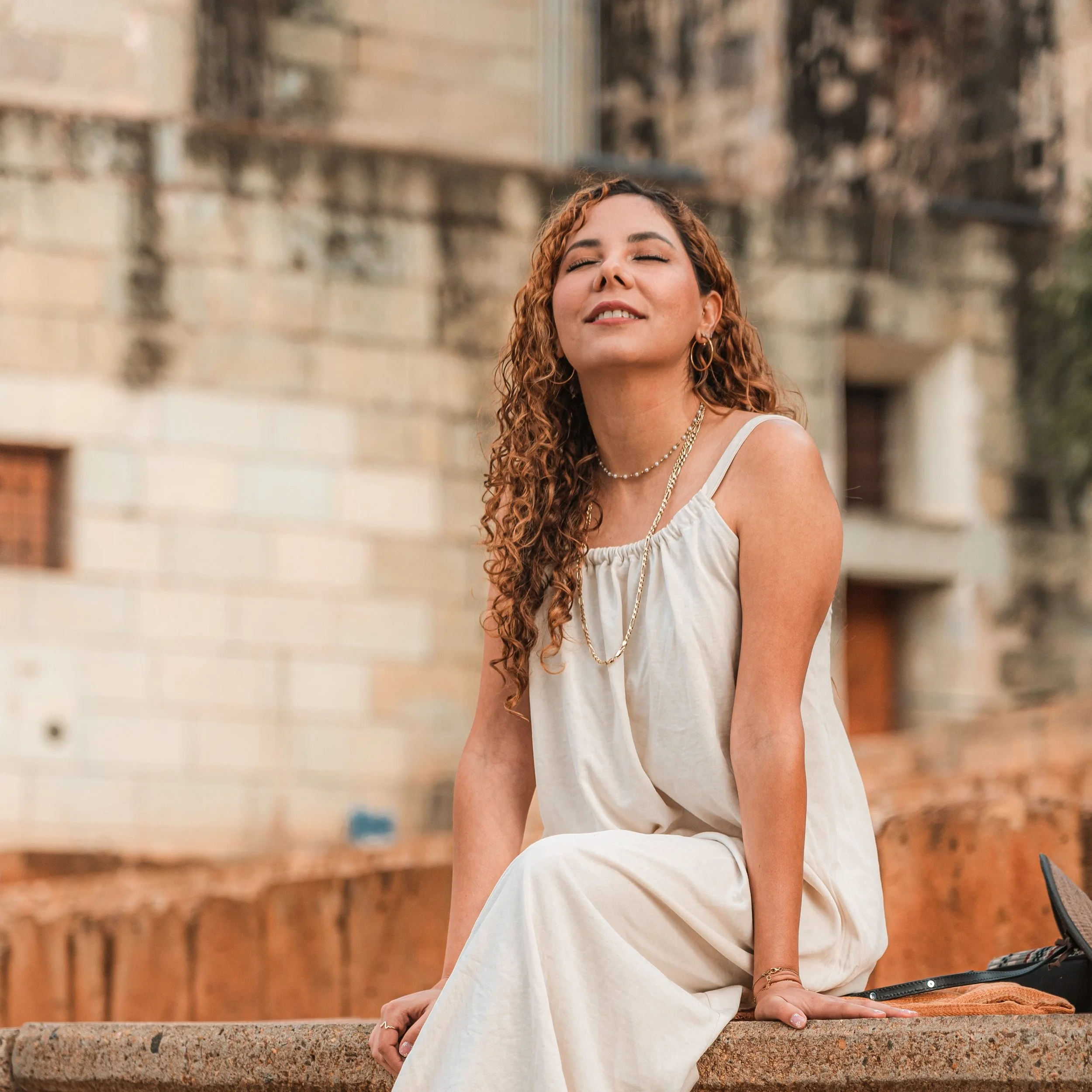 Mujer con vestido blanco sentada al aire libre, con edificios antiguos de fondo.