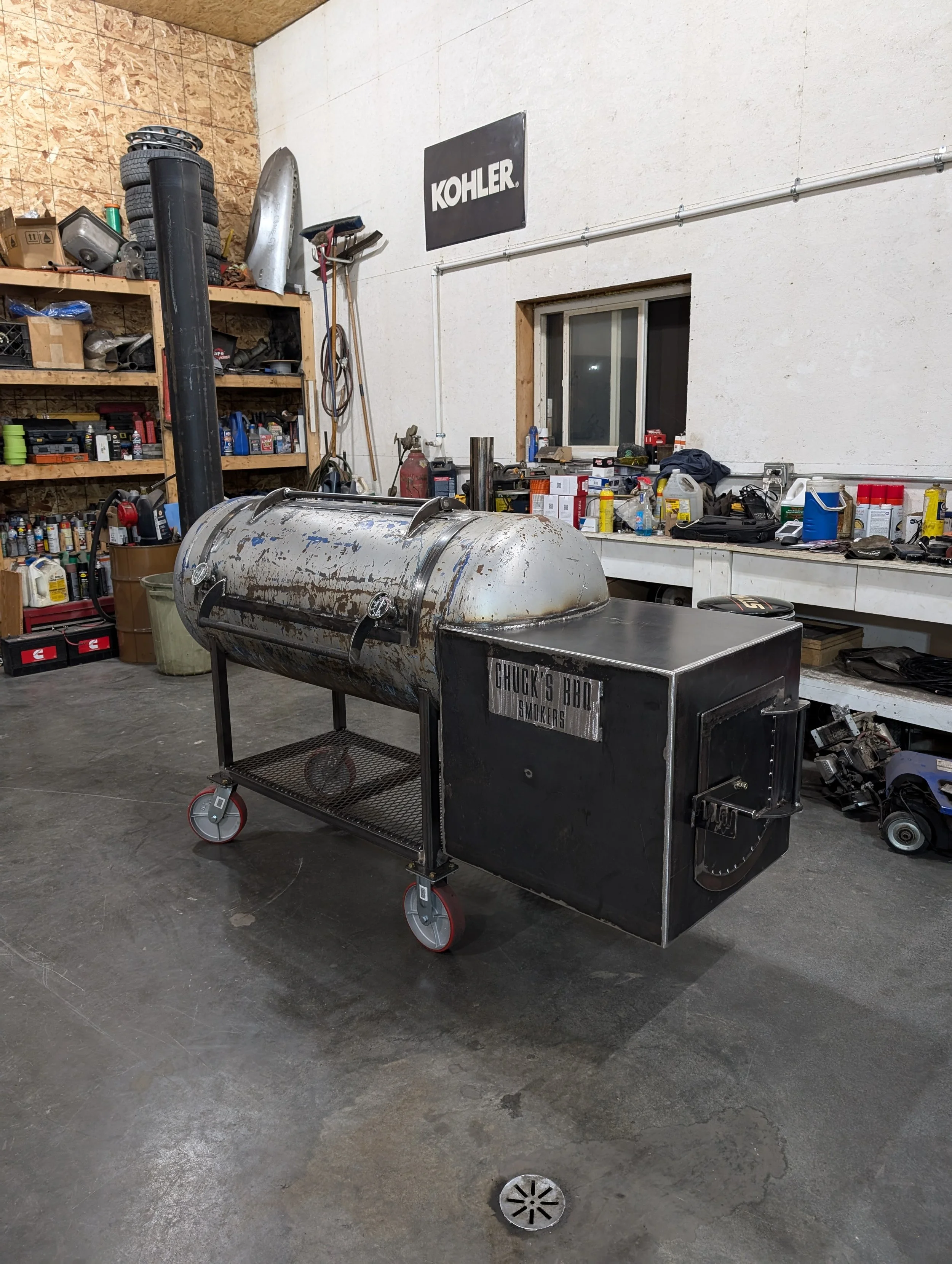 A smoker grill with a metal barrel smoking chamber and a black firebox attached, labeled 'Chuck's BBQ Smokers,' in a workshop setting with shelves and tools in the background.