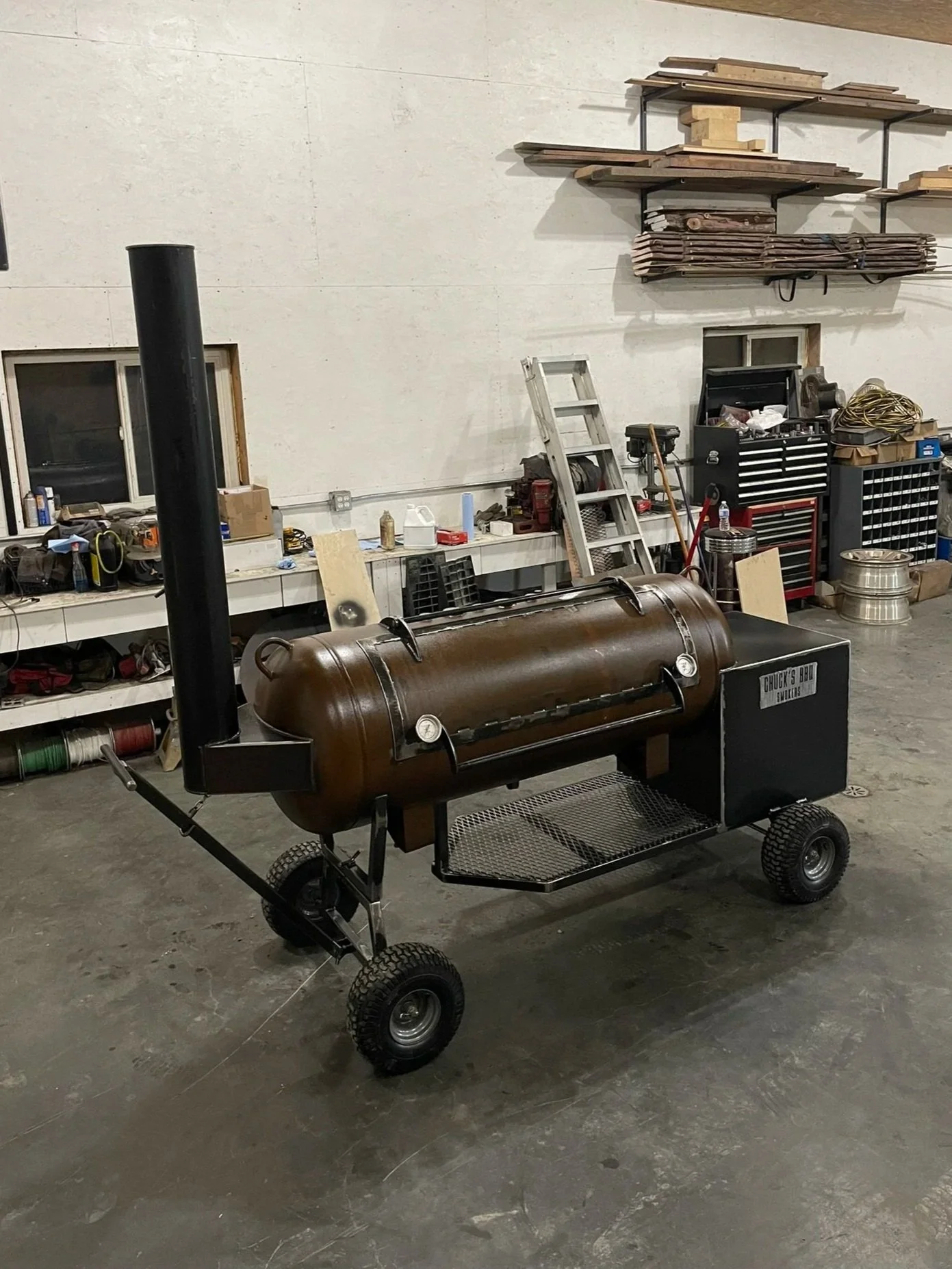 A smoker grill with a cylindrical metal chamber, black chimney, and wooden handle in an industrial workshop setting