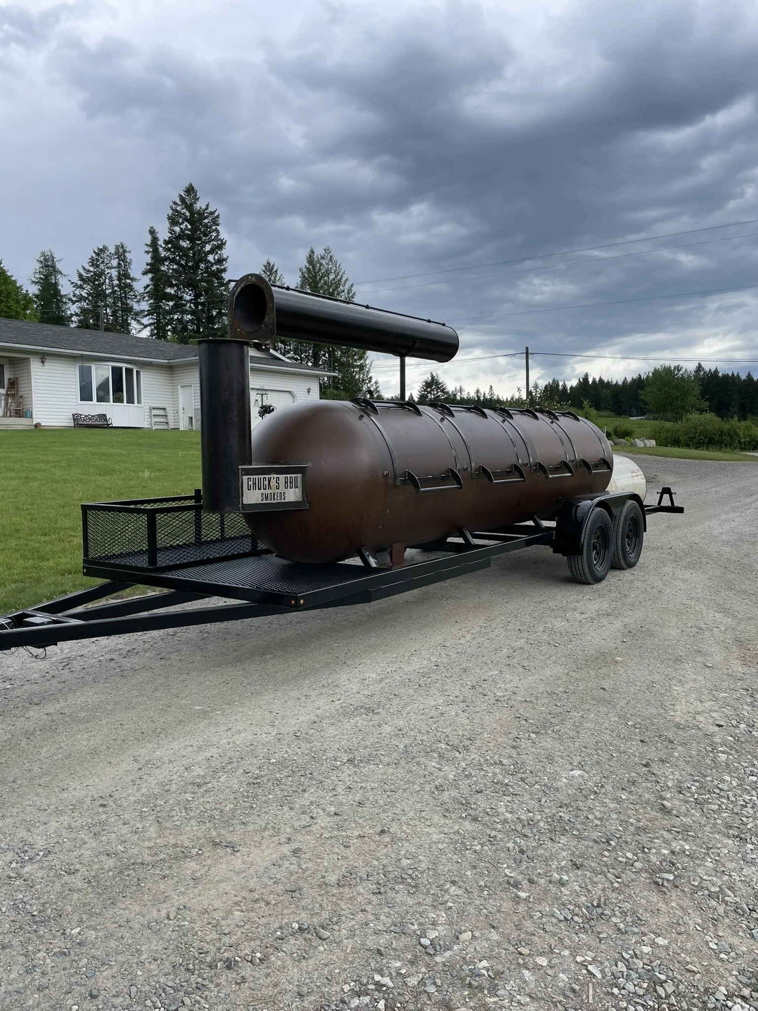A large cylindrical smoker on a trailer, labeled Chuck's BBQ Smokers, is parked on a gravel driveway outside with houses and trees visible in the background under a cloudy sky.