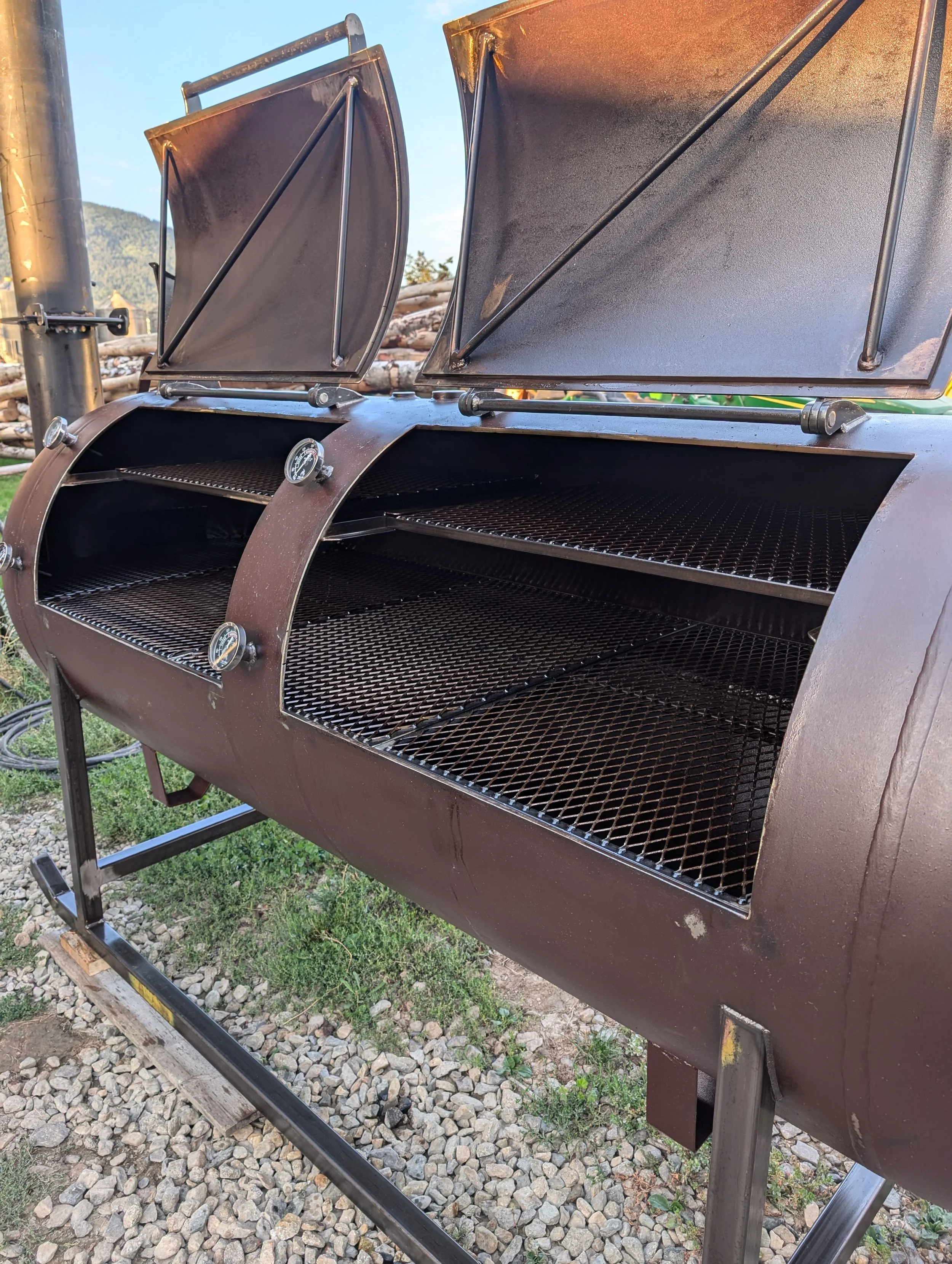 A large outdoor smoker with two metal lids open, showing multiple racks inside for smoking food. The smoker is on metal legs, with a thermometer on the side and a gravel ground underneath. Mountain scenery is visible in the background.