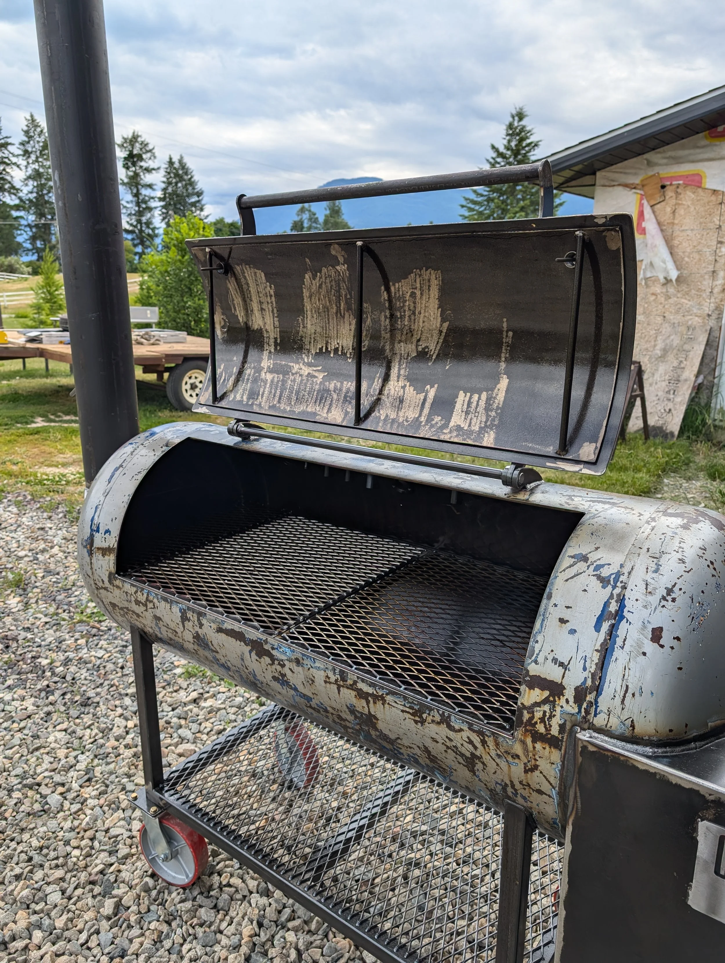 Patina smoker grill with an open lid, situated on a gravel surface with a rural background featuring trees and a cloudy sky.