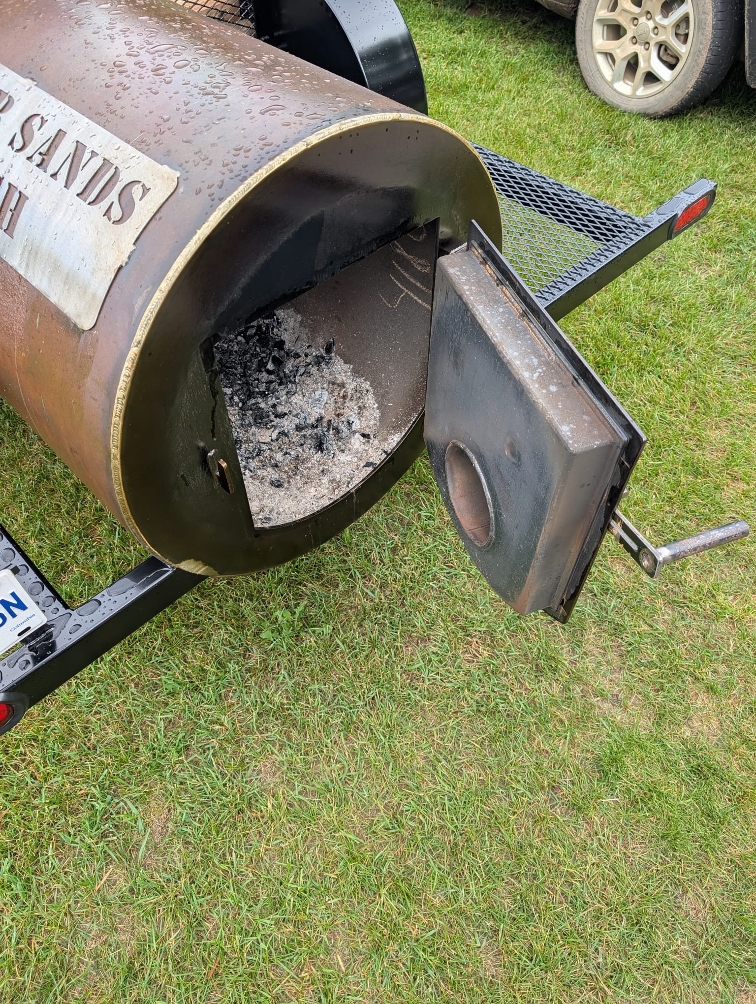 A large smoker firebox with its door open, showing burned wood inside. The barrel is mounted on a black metal trailer. There is grass on the ground and a vehicle tire in the background.