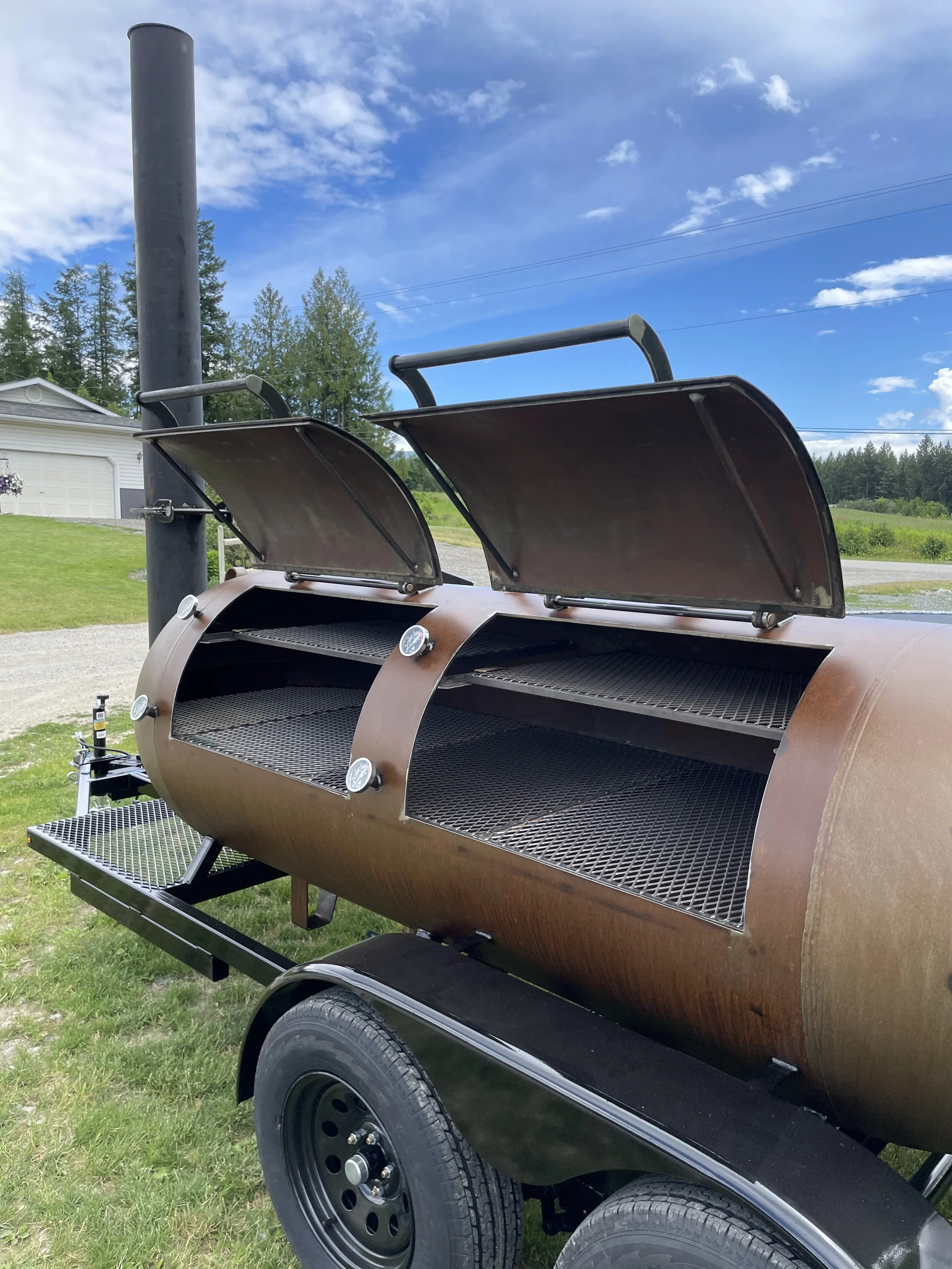 A large, outdoor BBQ smoker grill with two open lid compartments, a chimney pipe on the left, and a temperature gauge on the front, situated on a trailer in a grassy area with houses and trees in the background, under a partly cloudy sky.