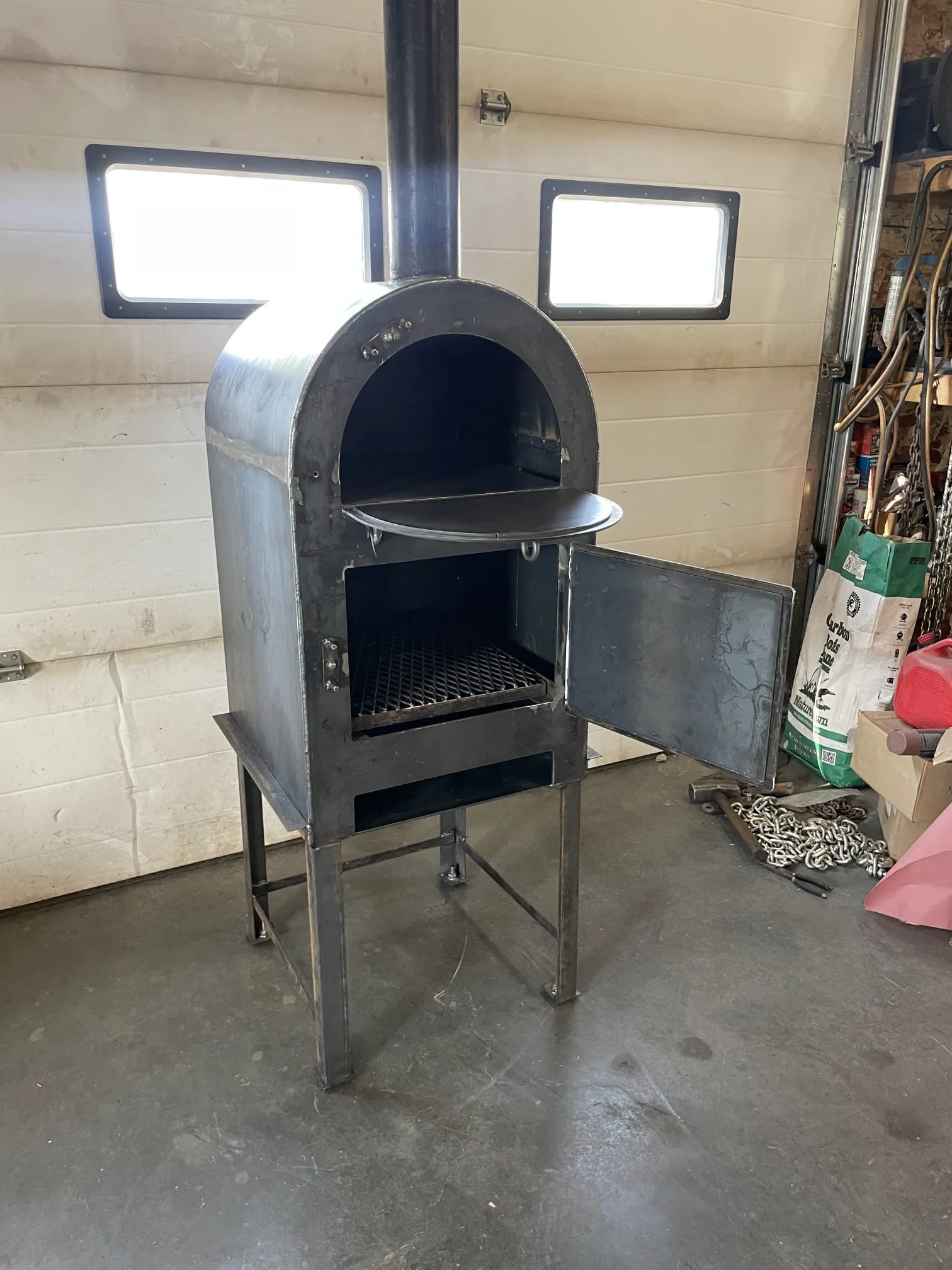 A stainless steel outdoor pizza oven with a chimney and a door open, revealing a grill and a pizza stone inside, in a garage or workshop setting.