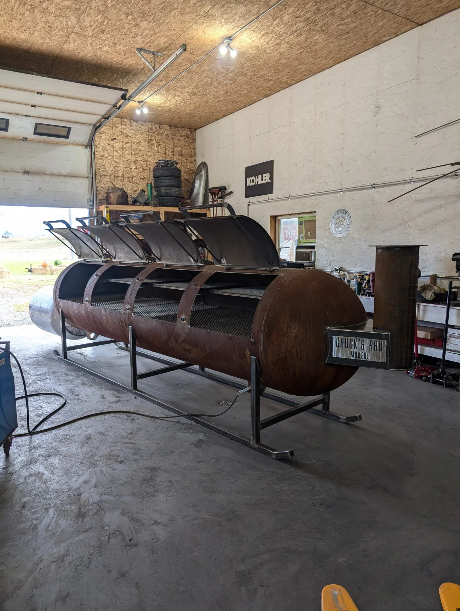 A large custom-made smoker with a rusty cylindrical body and multiple doors on top inside a workshop. The smoker has a metal stand and a plate that says 'Chuck's BBQ Smokers'.