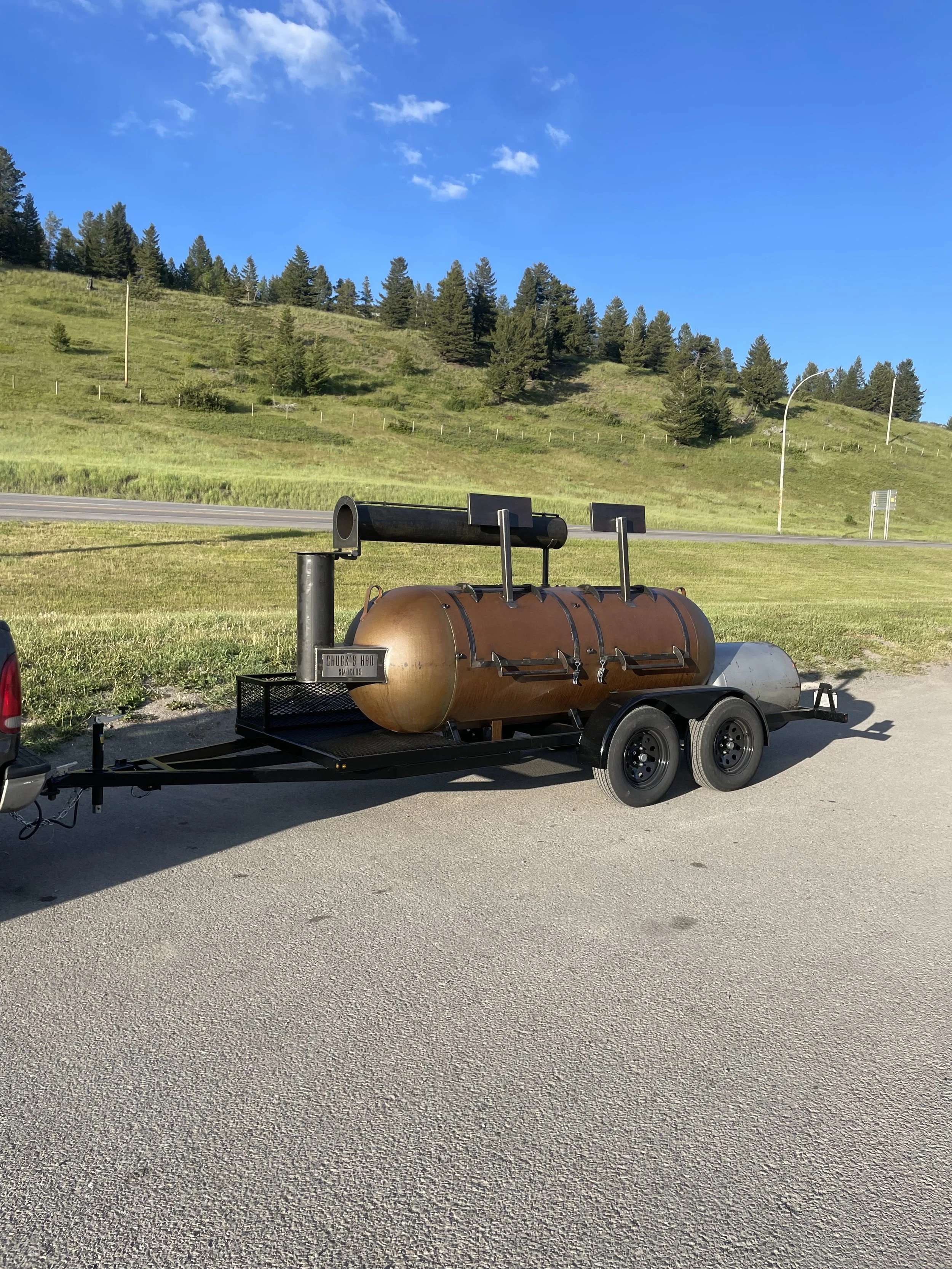 A large barbecue smoker on wheels attached to a vehicle, set on a paved parking lot with grassy hills and trees in the background under a blue sky.