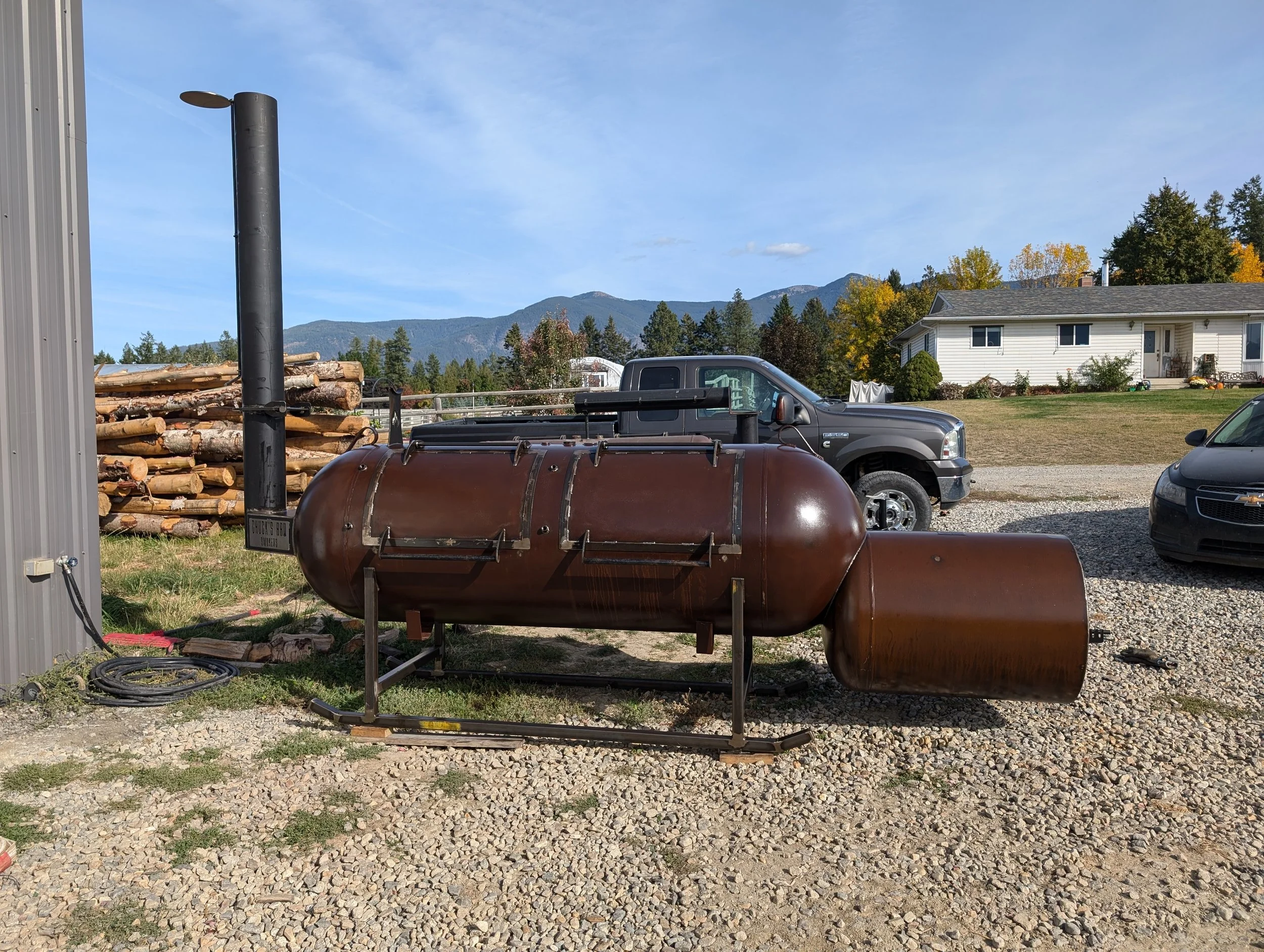 A rusty colored smoker grill on a patch of grass and gravel with a black truck, a house, and mountains in the background.