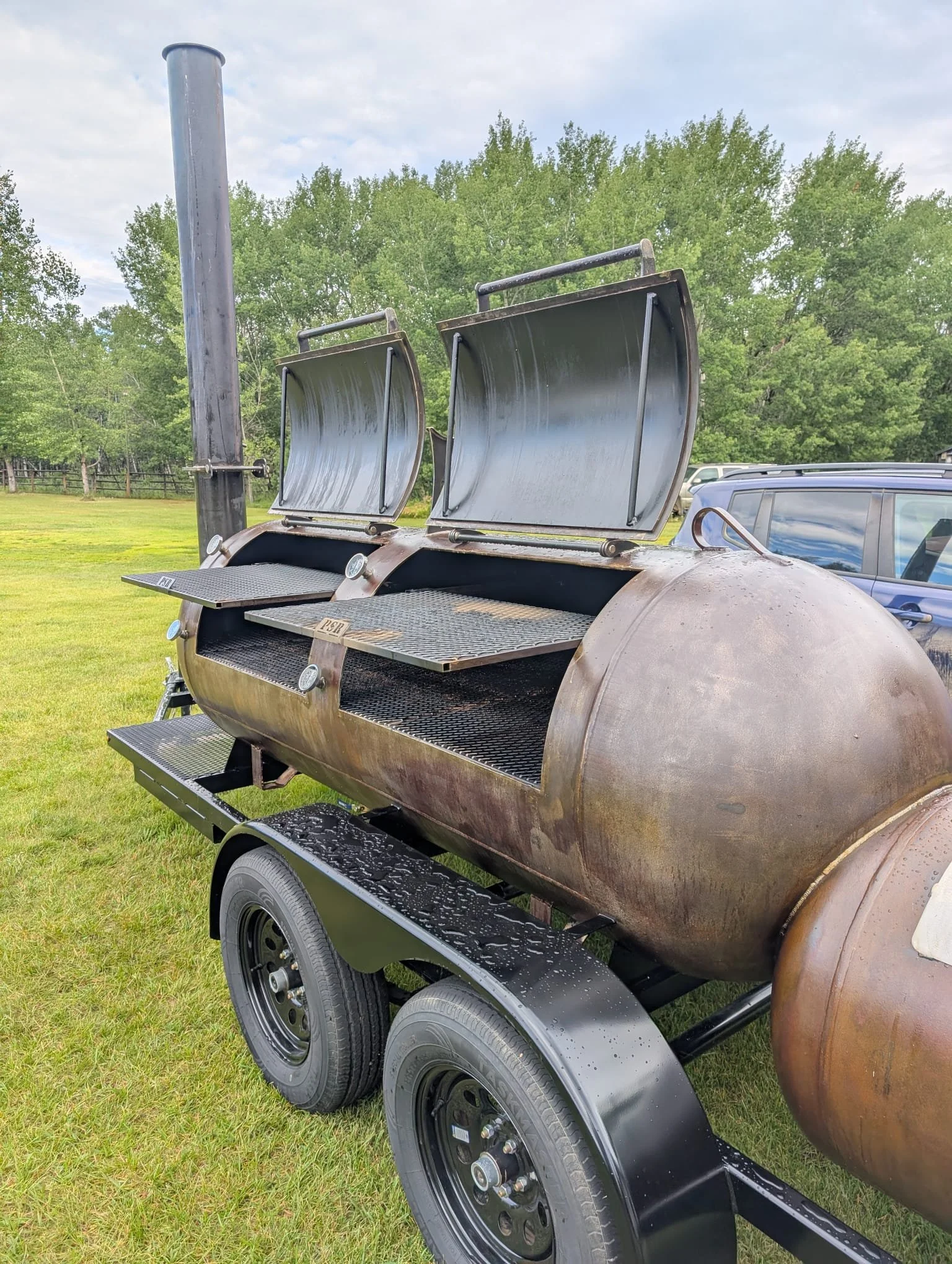 A large, rust colored smoker grill on a black trailer with two wheels, situated outdoors on grass with trees and a partly cloudy sky in the background.