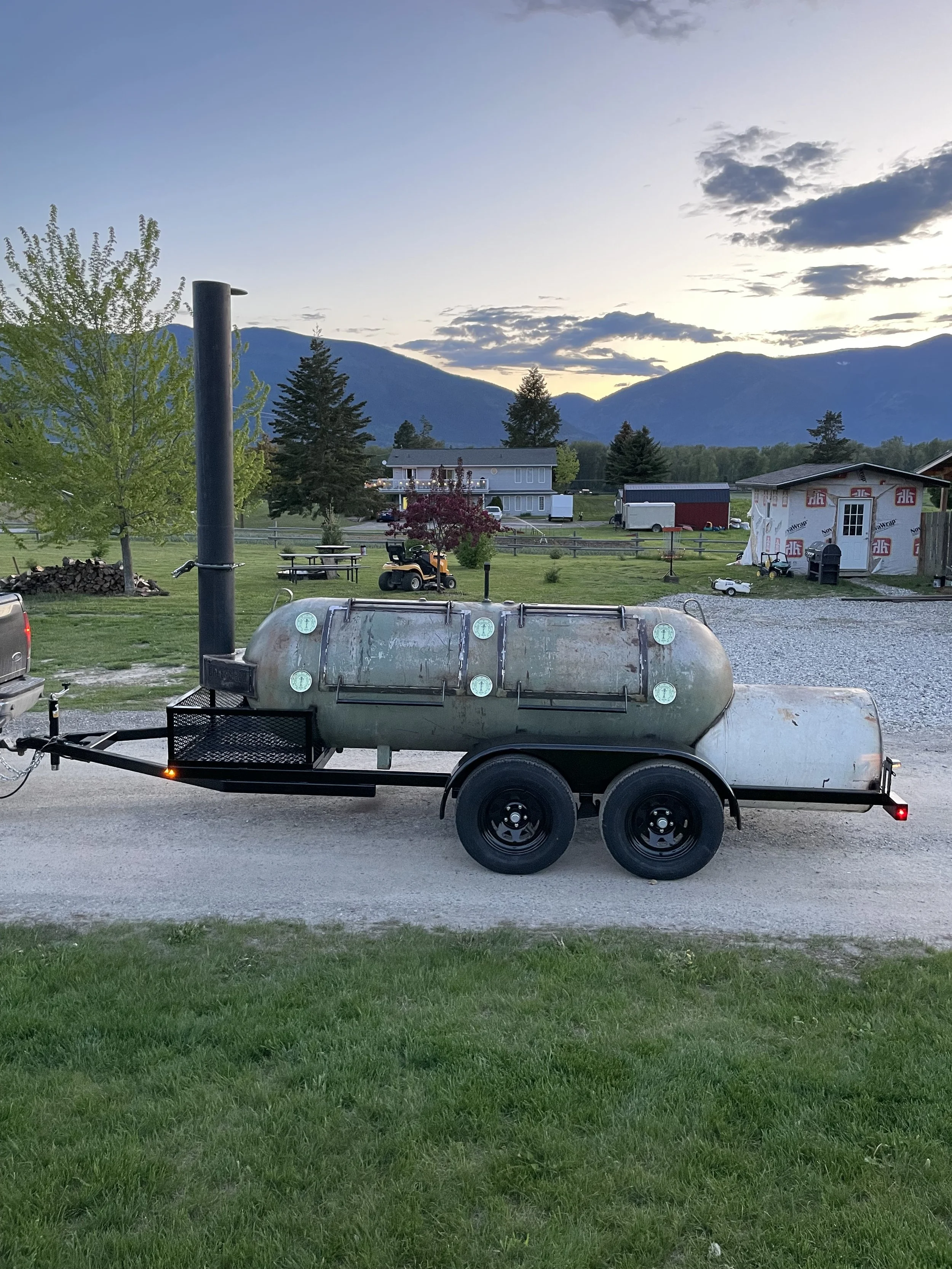 A metal barbecue smoker on a trailer parked on a gravel driveway with green grass in the foreground, against a rural background of trees, houses, a mountain range, and a cloudy sky at sunset.