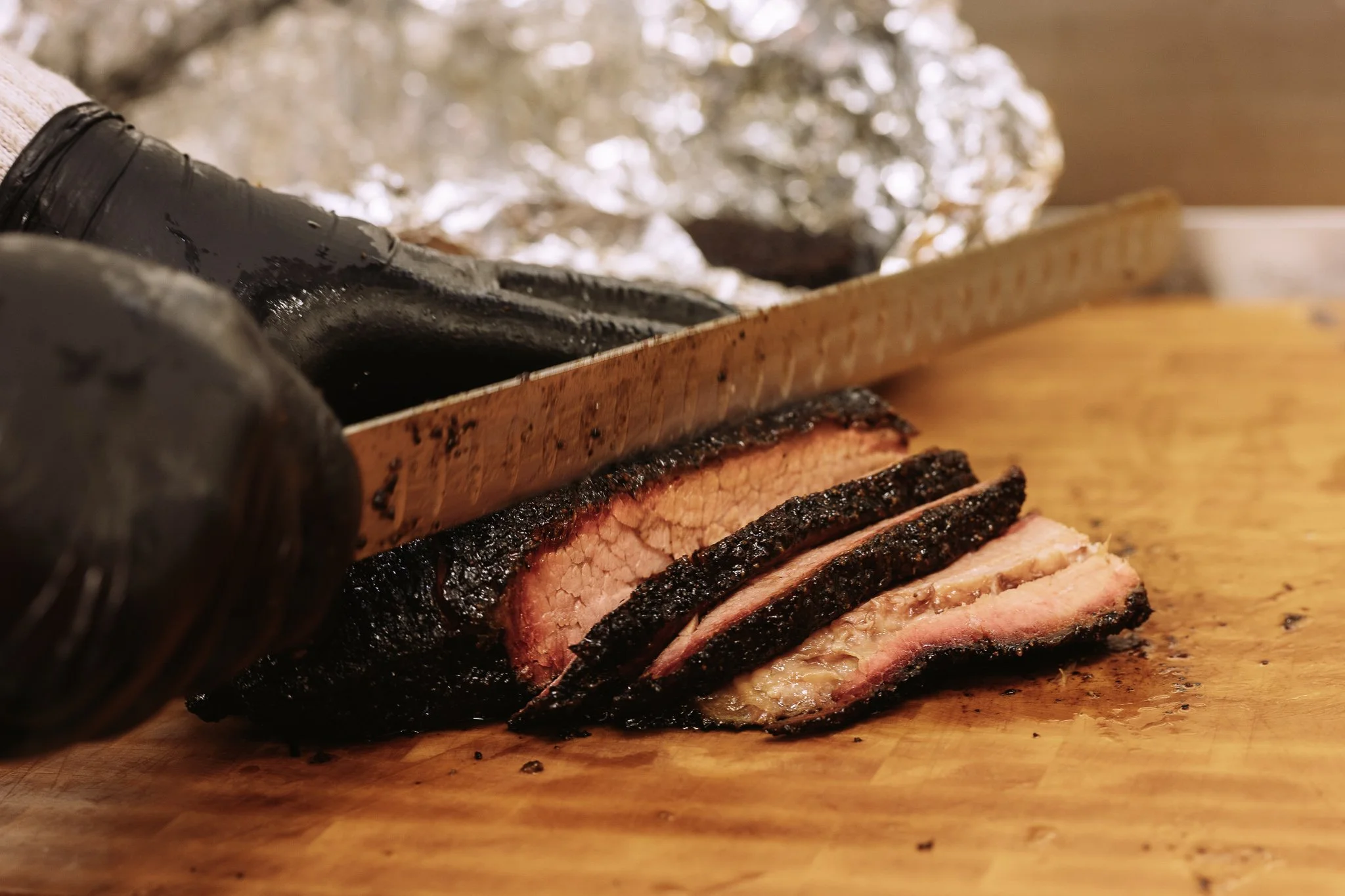 Sliced smoked brisket on a wooden cutting board with foil-wrapped food in the background.