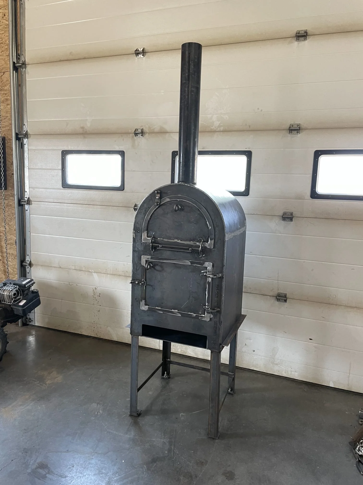 A metal outdoor pizza oven with a chimney pipe, standing on a metal frame in a garage with a white garage door and small windows.