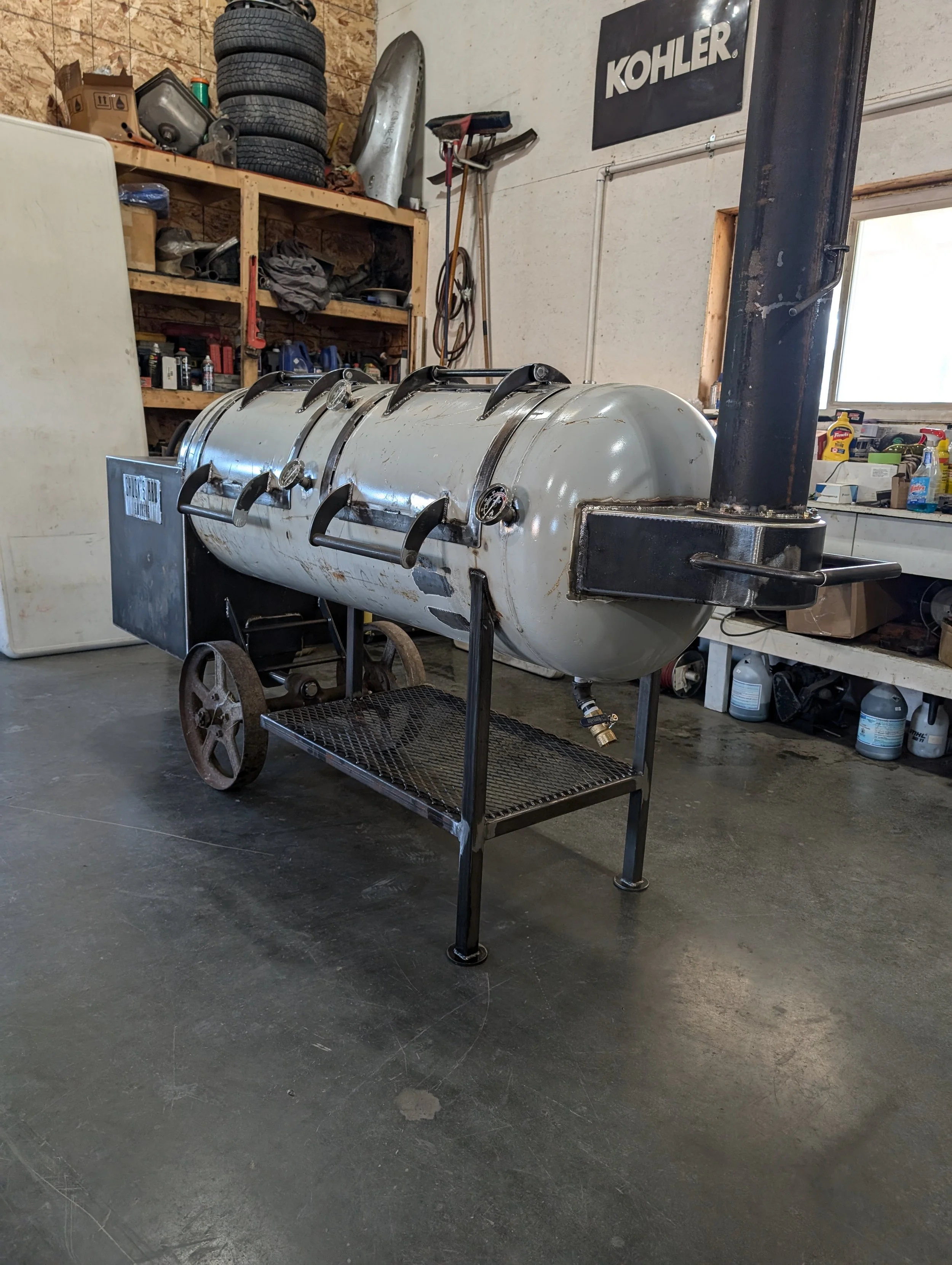 A smoker or barbecue grill with a cylindrical body on wheels, located in a garage workshop with shelves and tools in the background.
