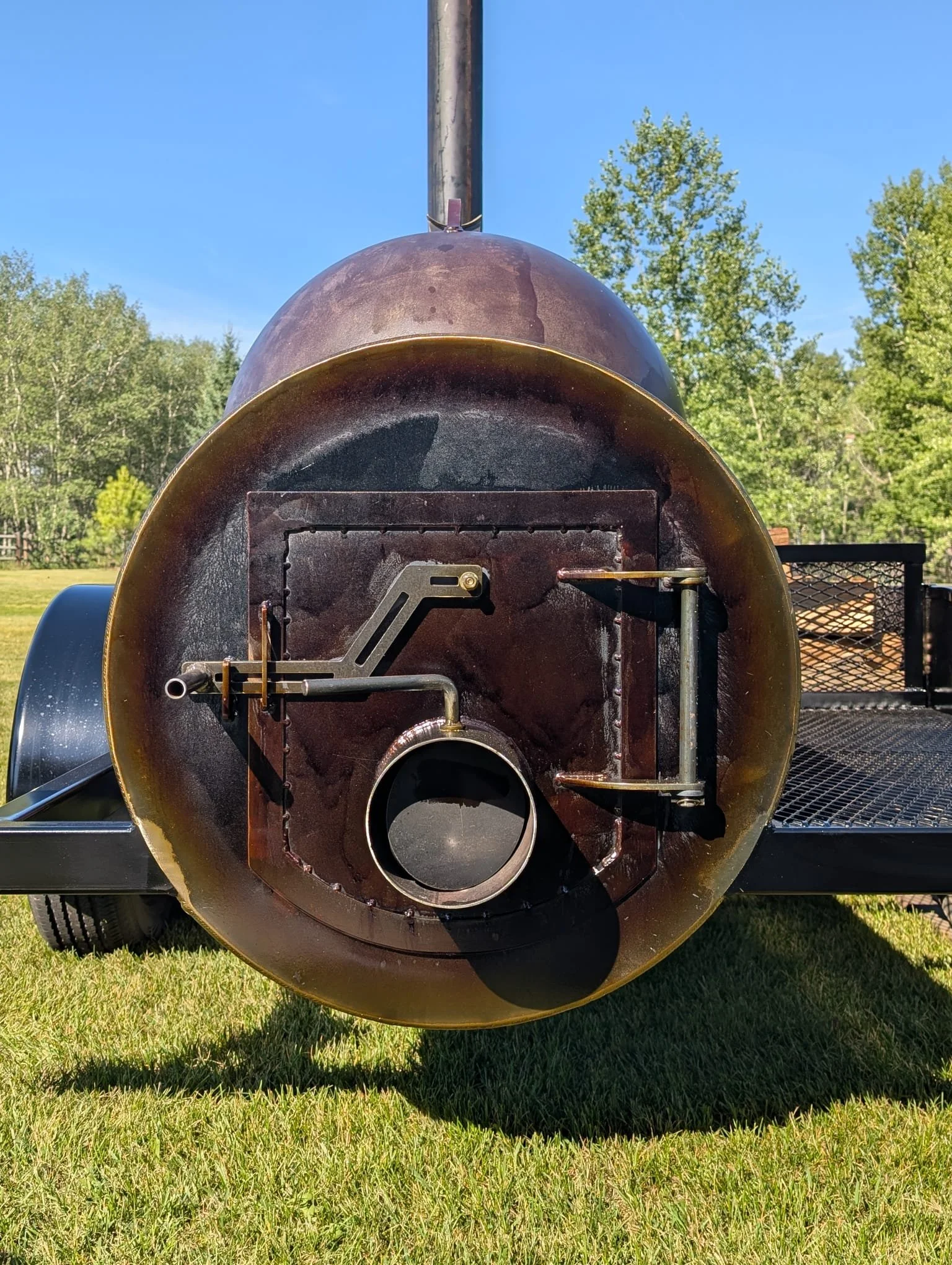 Back view of a large metal smoker grill on a trailer, with a grassy field and trees in the background.