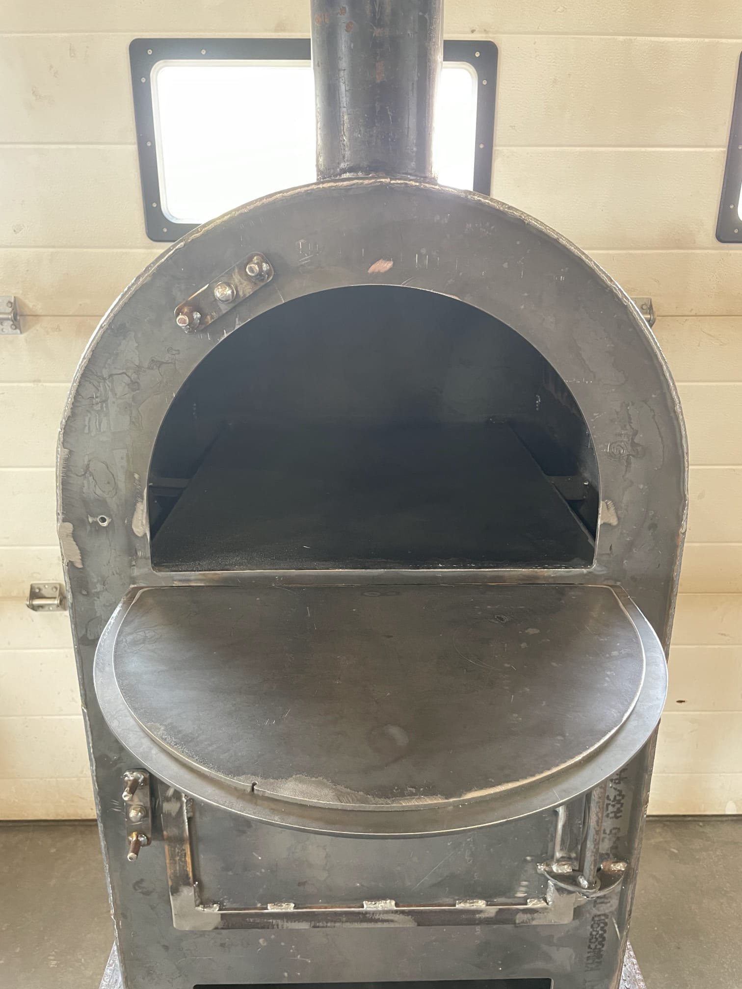 Empty stainless steel pizza oven with a rounded opening and a shelf inside, against a beige wall.