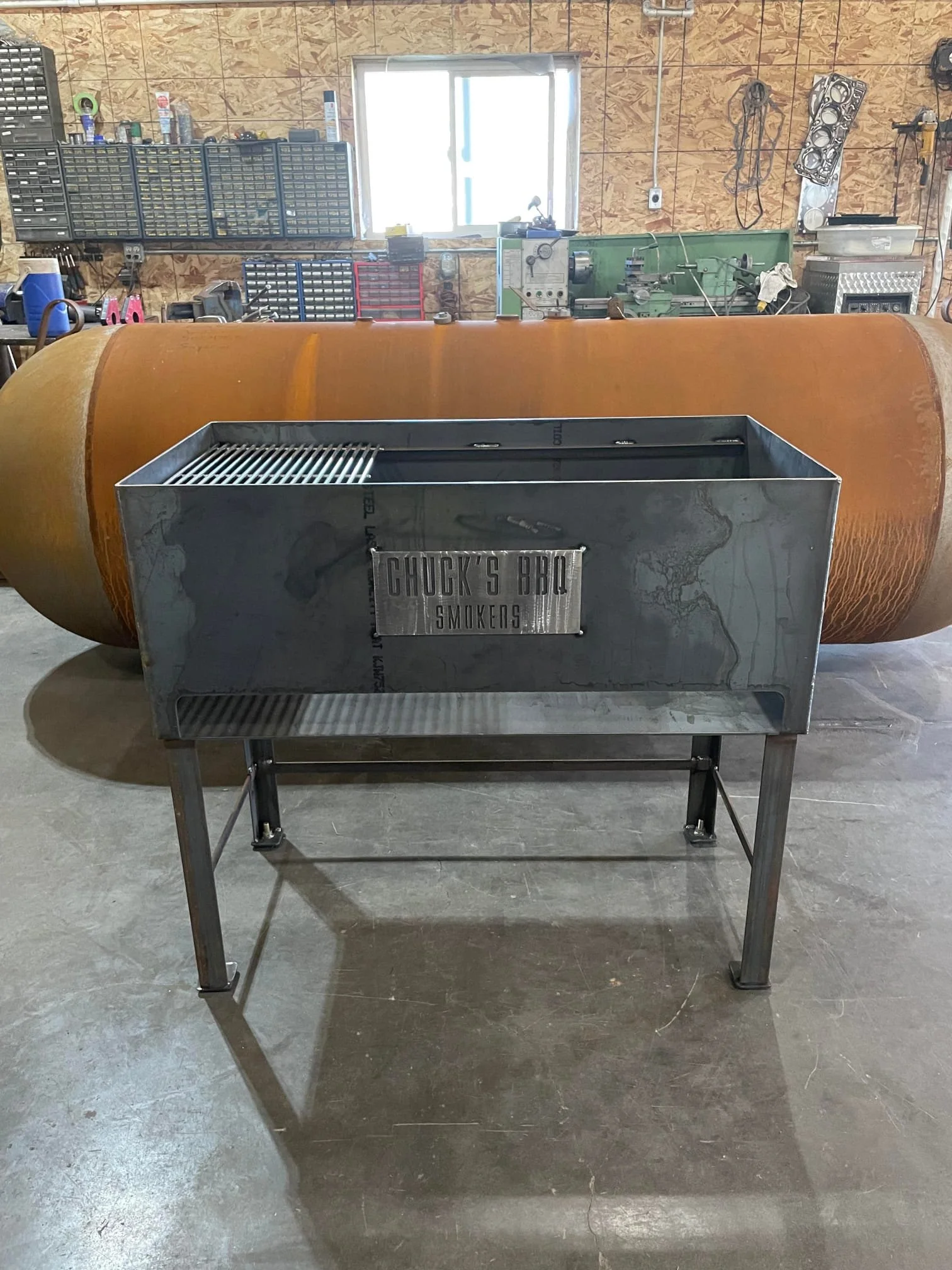 A metal grill with a sign that reads 'Chuck's BBQ Smokes' on the front, placed in front of a large rusted propane tank in a workshop with various tools and equipment on shelves and walls.