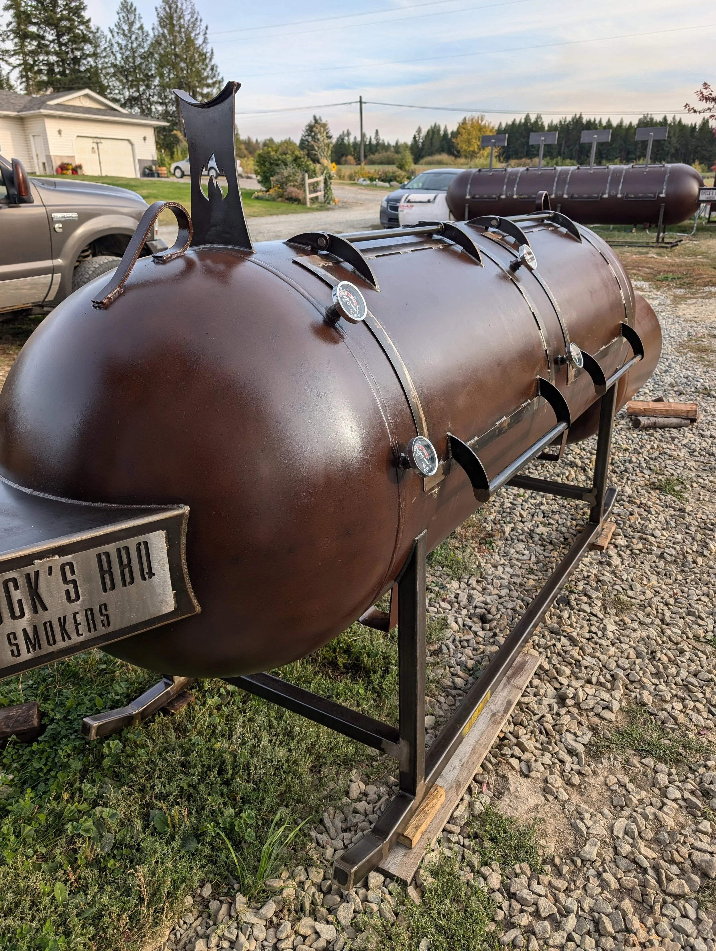 Large rust-colored barrel-style smoker grill with thermometer gauges and a sign that reads 'Chuck's BBQ Smokers', set outdoors on a gravel surface with grass and trees in the background.