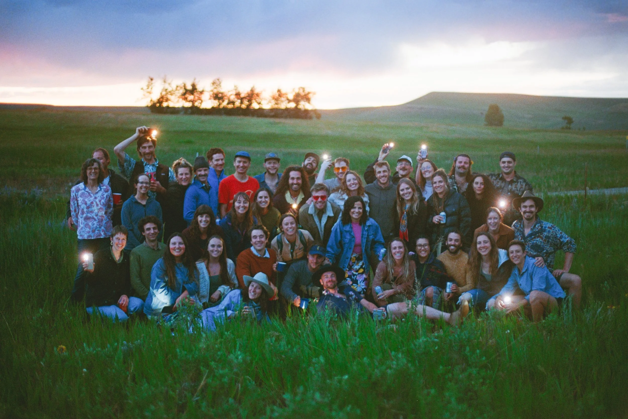 Outdoor film group portrait during a Montana wedding weekend welcome party the night before the wedding, photographed at sunset in an open field with phone lights.