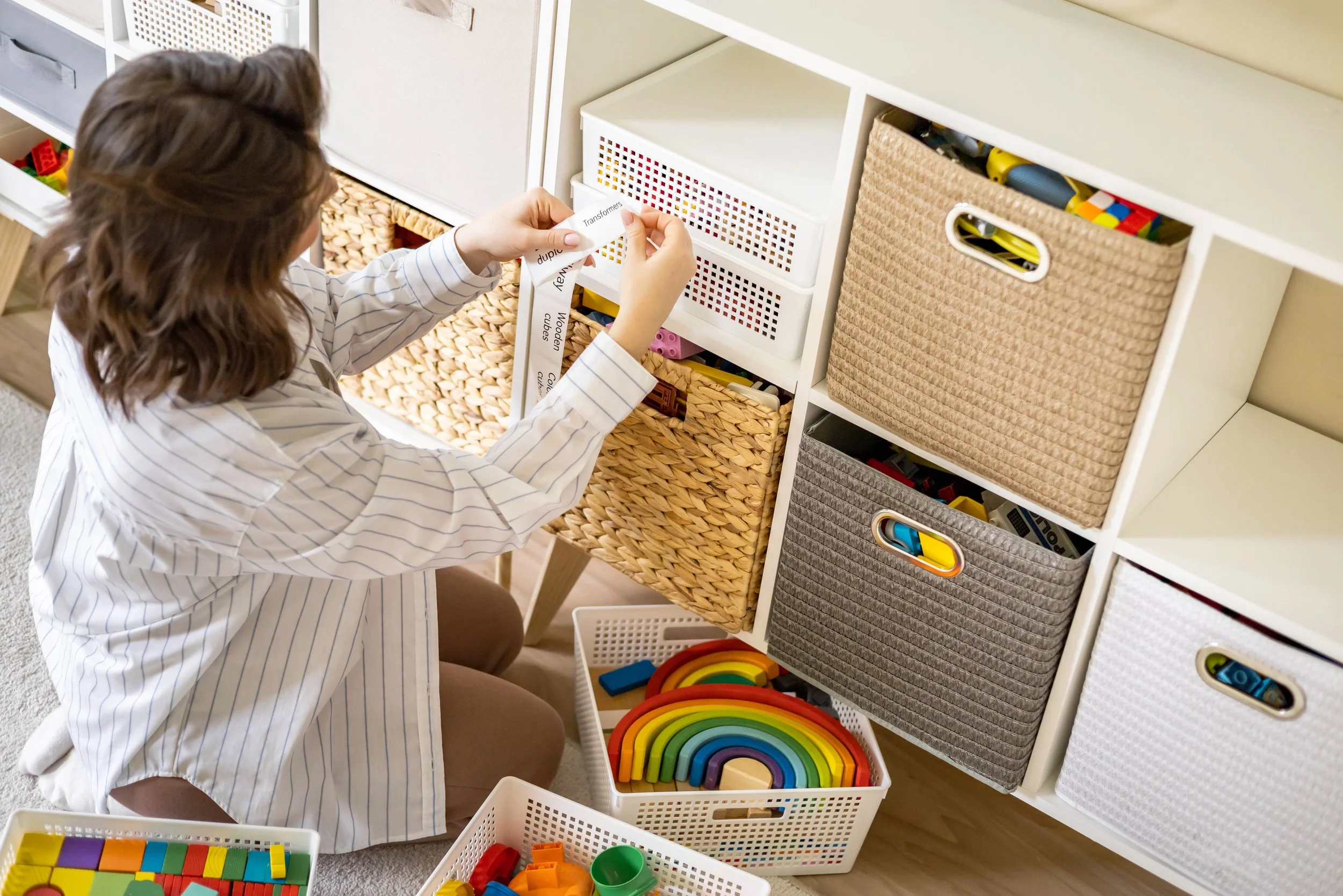 A woman organizing toys in a white storage unit with fabric baskets and open shelves, surrounded by toy baskets containing colorful building blocks and rainbow-shaped toys.