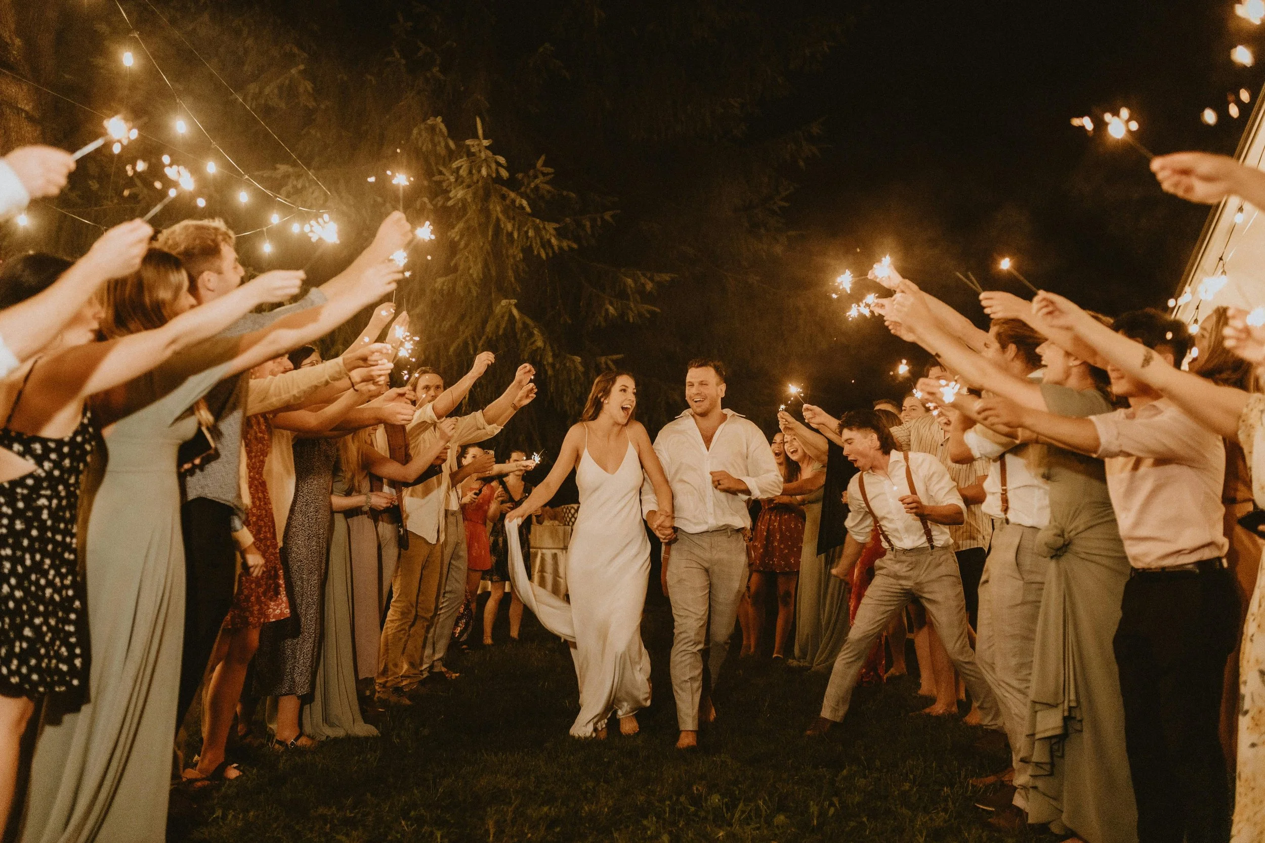 A bride and groom walking hand in hand through a crowd of friends at night, holding sparklers during a celebration or wedding reception.