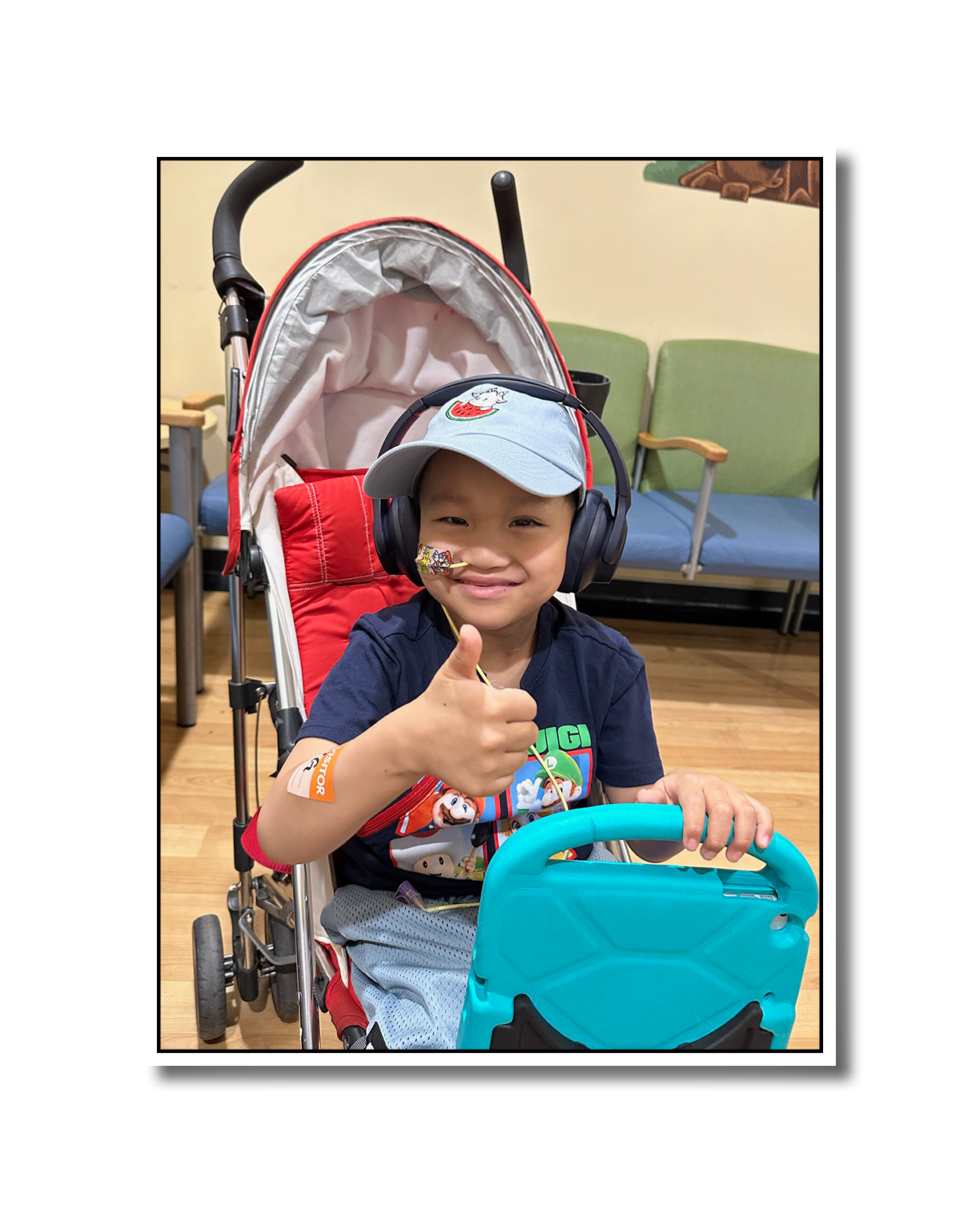 A young boy sitting in a stroller, smiling and giving a thumbs-up. He is wearing a baseball cap, large headphones, and has a nasal cannula. The background shows a waiting room with chairs.