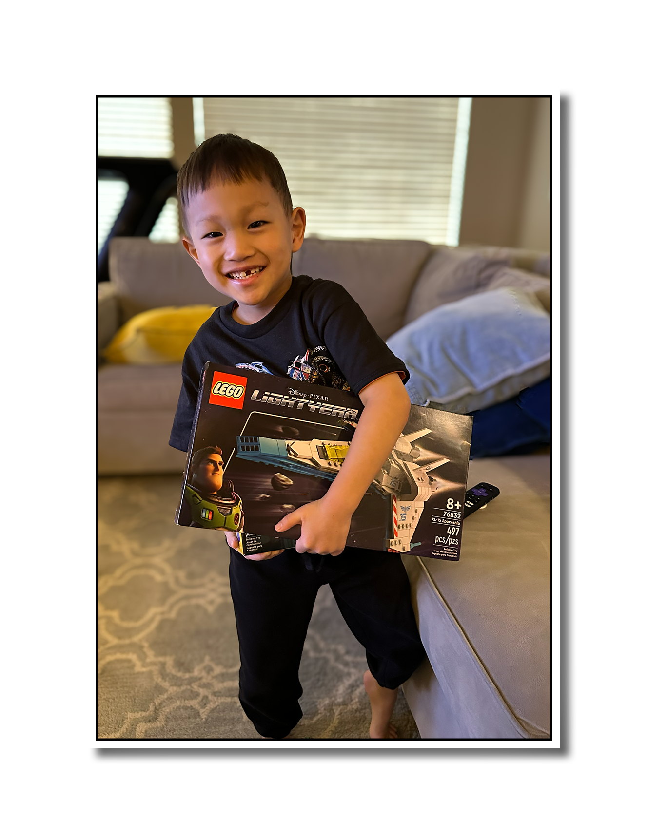A young boy standing in a living room, smiling and holding a LEGO Lightyear spaceship set box.